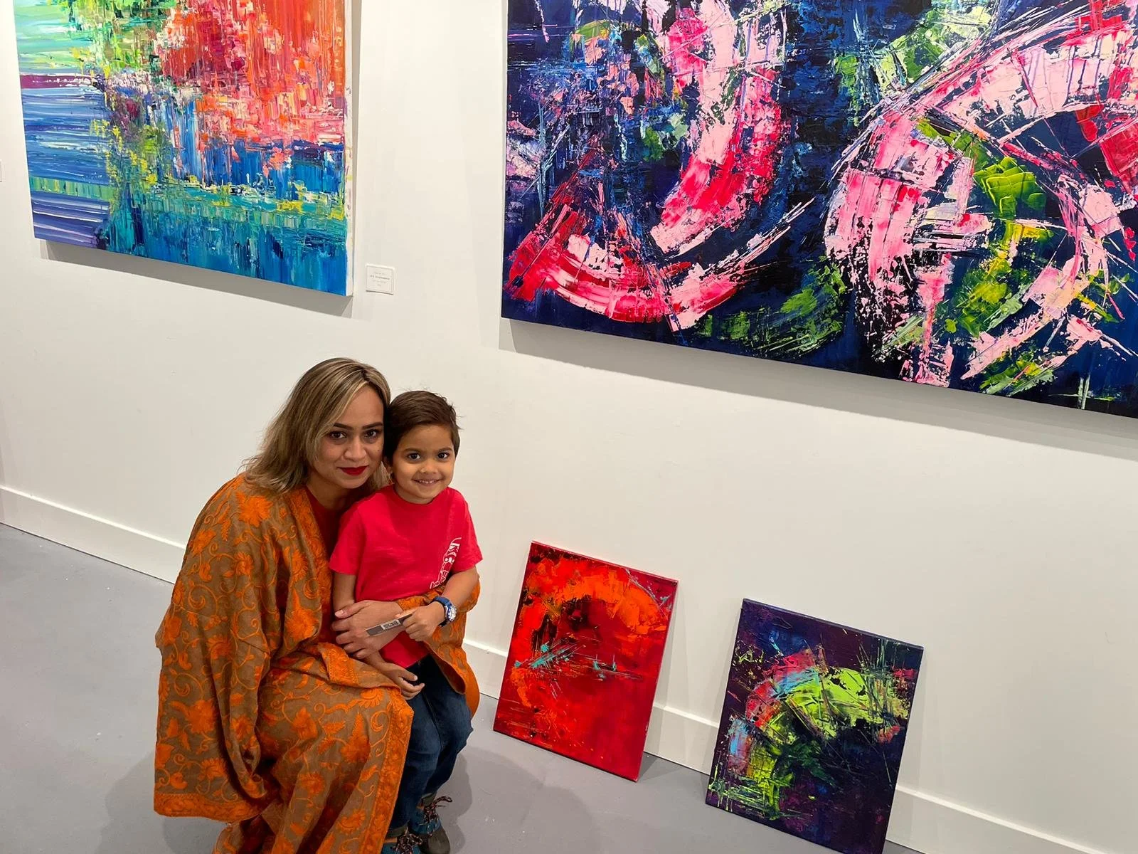 A woman in an orange dress and a young girl in a red shirt are sitting together in an art gallery, smiling at the camera. Behind them on the white wall are several colorful abstract paintings, with two more paintings resting on the floor.