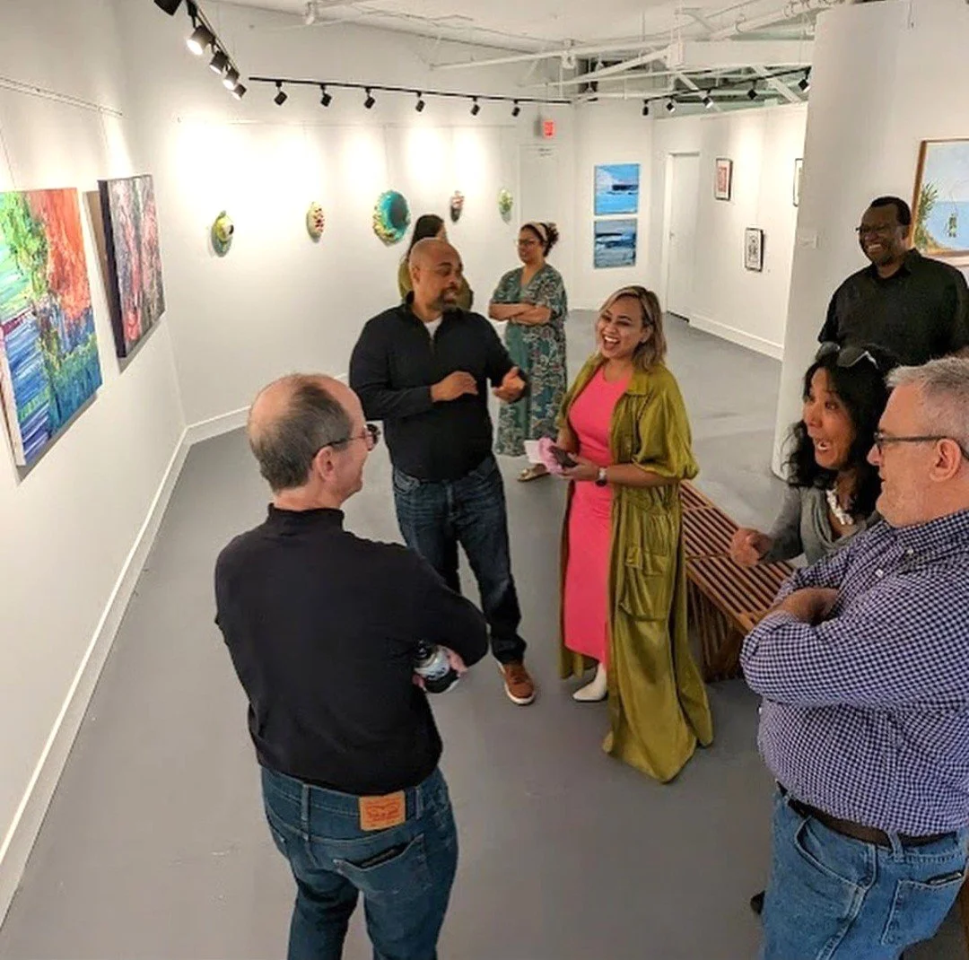 A group of people socializing at an art gallery, standing and sitting near colorful paintings and wall art.