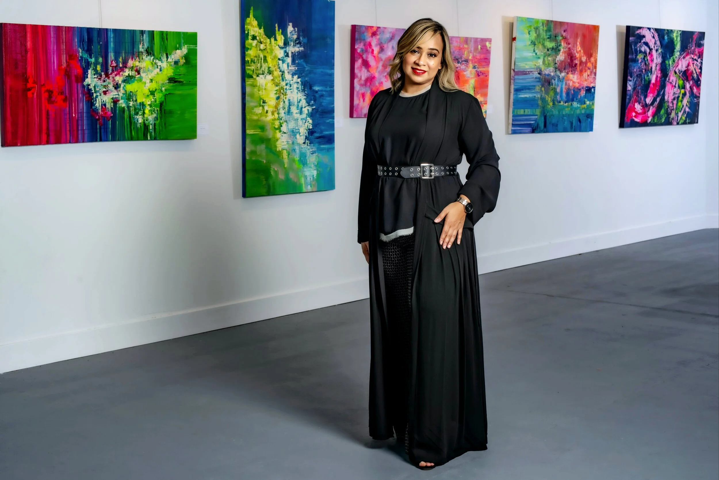 Woman in black dress standing in an art gallery with colorful abstract paintings on the wall.