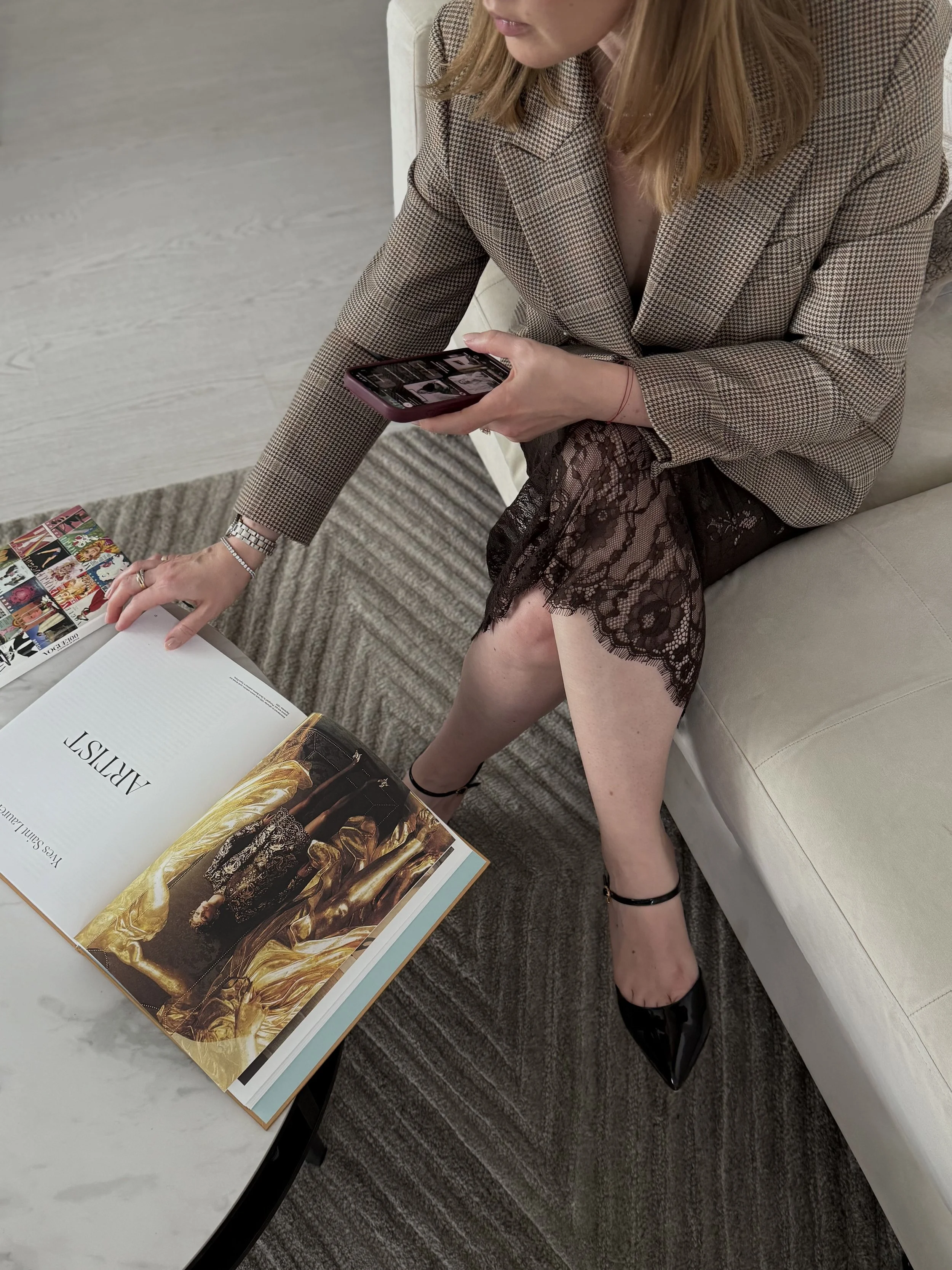 A woman sitting on a white couch, looking at her phone. She is wearing a plaid blazer, a black lace skirt, and black high heels. There is an open magazine on the table in front of her, and a sheet of paper with multiple images on it.
