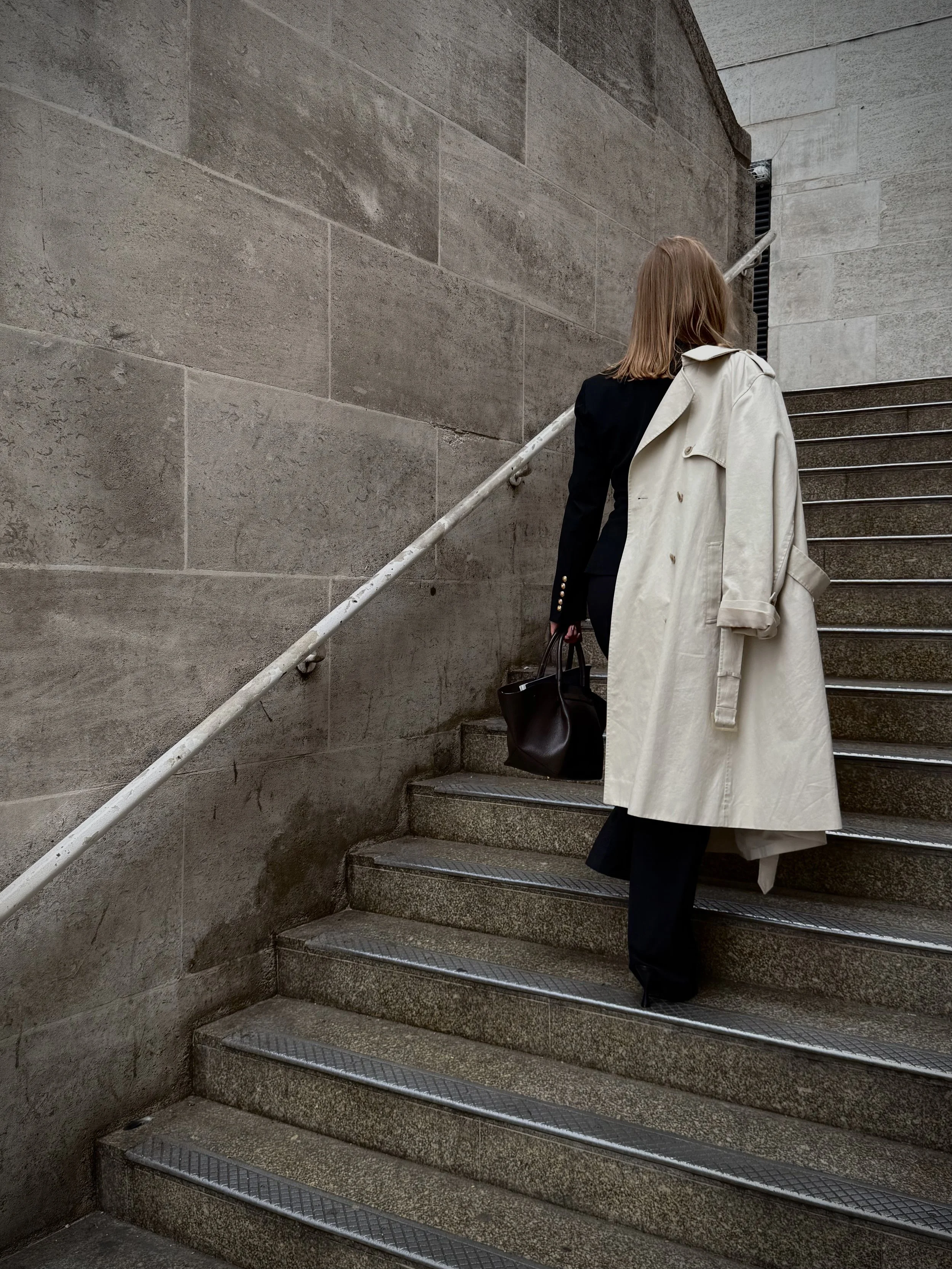 A woman with red hair walking up an outdoor stairway, carrying a black handbag and wearing a beige trench coat draped over her shoulders, with a black blazer and black trousers underneath.