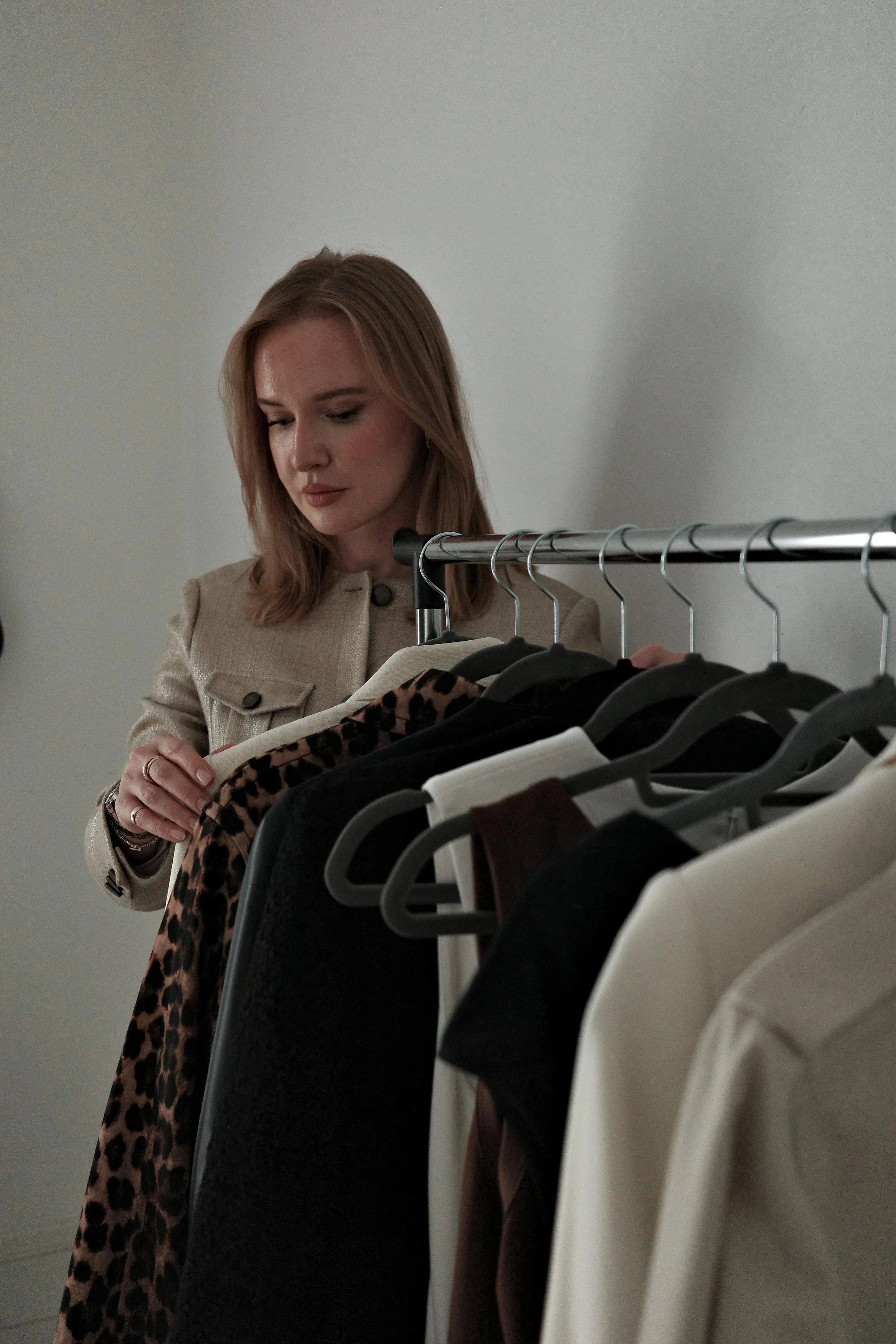 A woman with red hair looking at clothing on a rack, holding a beige coat, in a minimalistic setting.