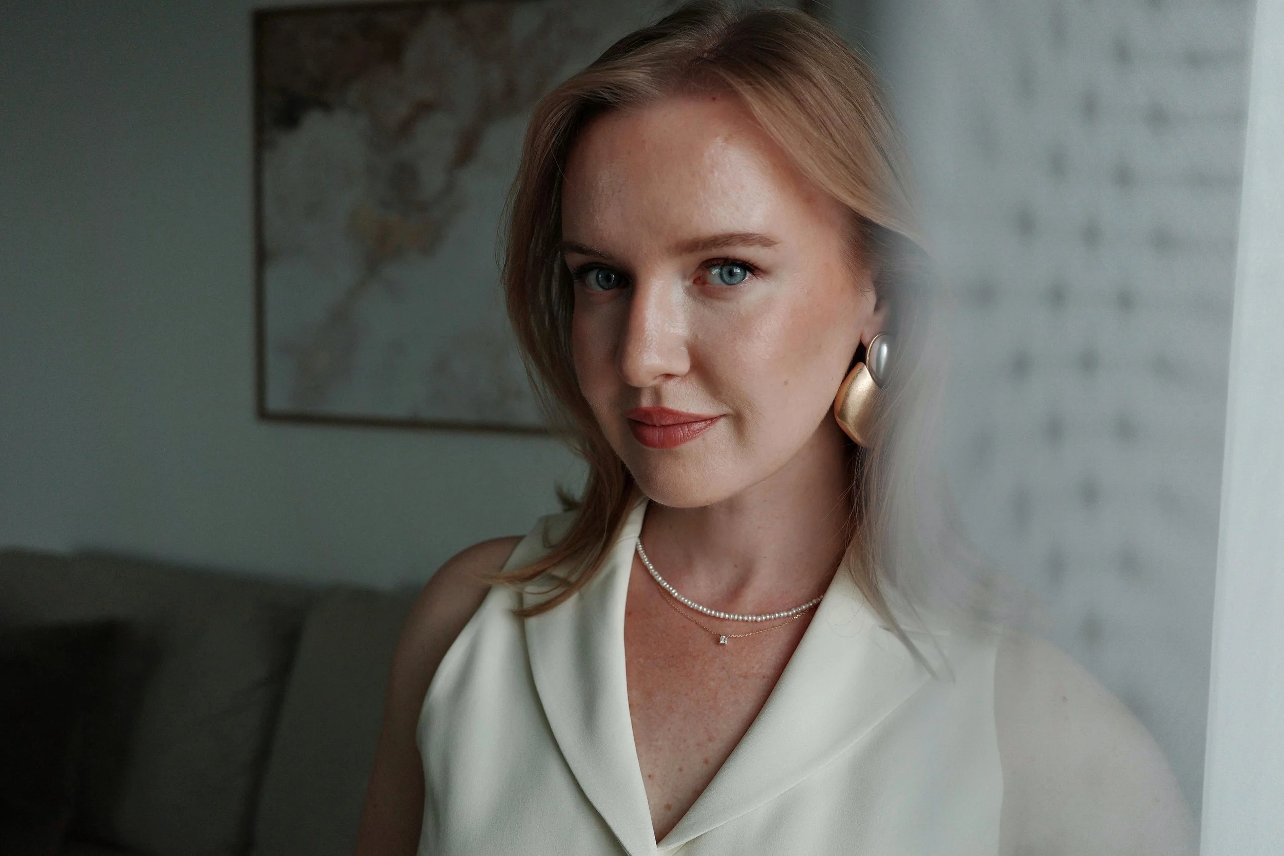 A woman with shoulder-length blonde hair, blue eyes, wearing a cream sleeveless top, pearl necklace, and large earrings, standing indoors near a window with a blurred map in the background.