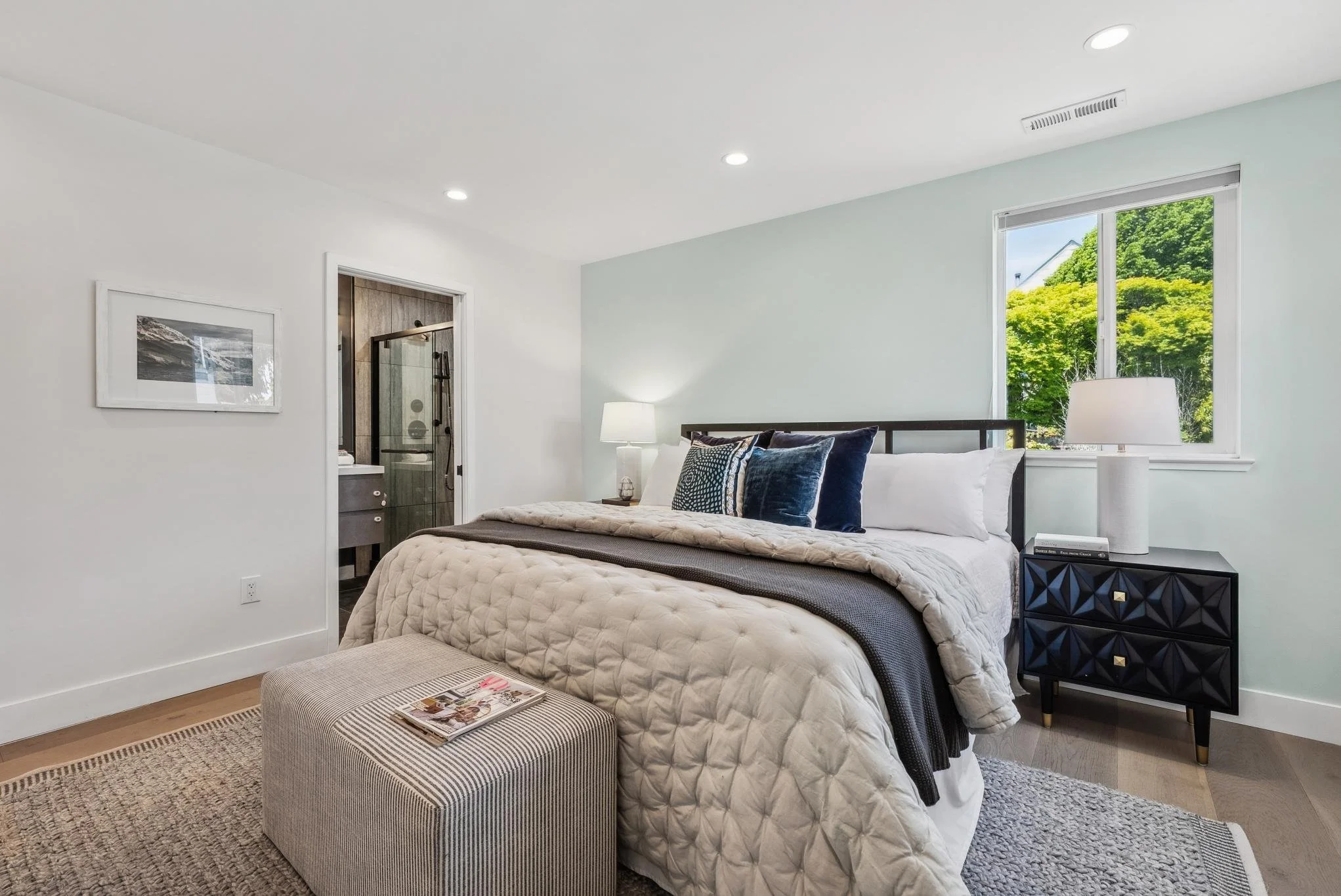 Modern bedroom with a neatly made bed featuring a quilted comforter and decorative pillows. A nightstand with a lamp sits beside the bed. A bathroom is visible through an open door. A window with a view of green trees illuminates the room. Artwork ha