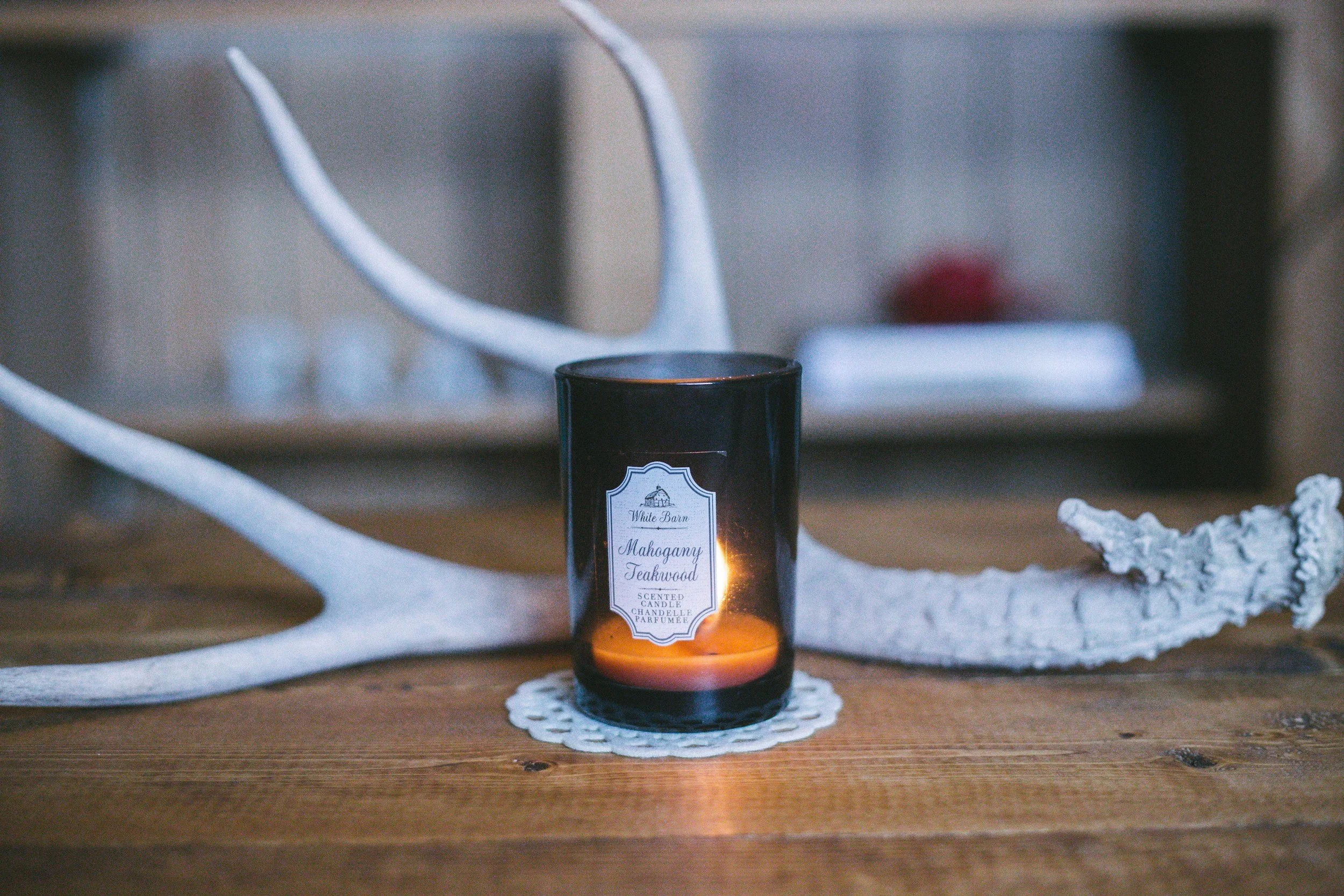 A black scented candle labeled 'Mahogany Teakwood' on a wooden table, with a large white antler behind it and a white lace doily underneath.