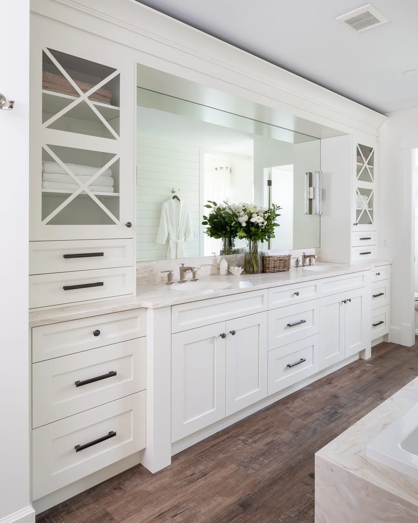 A luxurious white bathroom with a marble countertop, black handles on cabinets and drawers, a large mirror, and a vase with white flowers.
