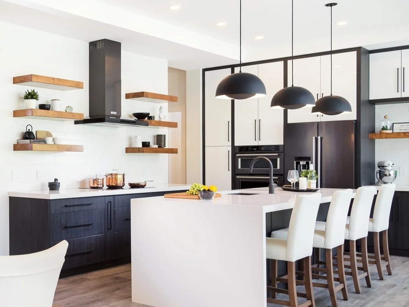 Modern kitchen with black and white cabinetry, a white island with four white chairs, open wooden shelves with plants and decorative items, black pendant lights above the island, and stainless steel appliances.