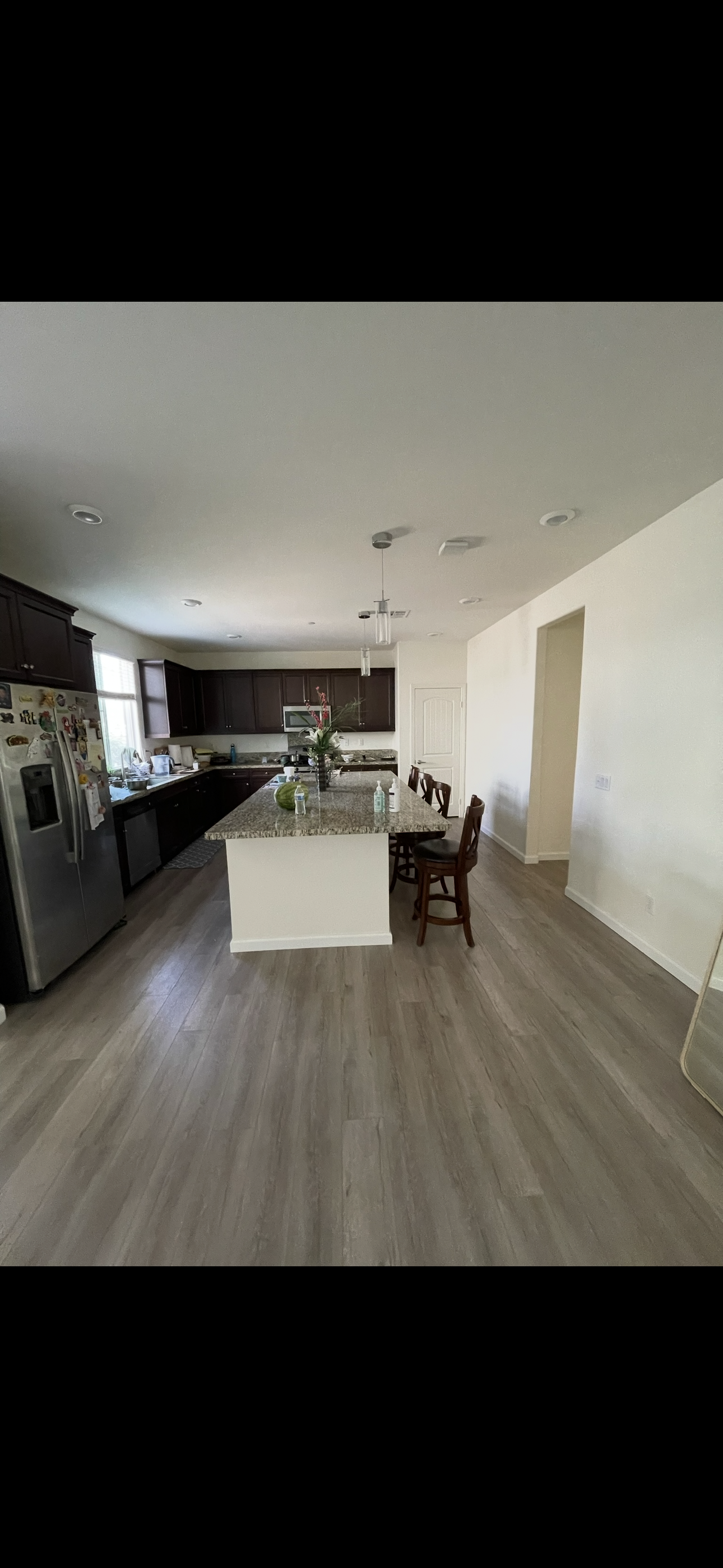 A modern kitchen with dark wood cabinets, stainless steel appliances, a large granite island with a flower arrangement, and barstools. Light wood flooring, white walls, and a small window with sunlight. Various kitchen items on the countertops.