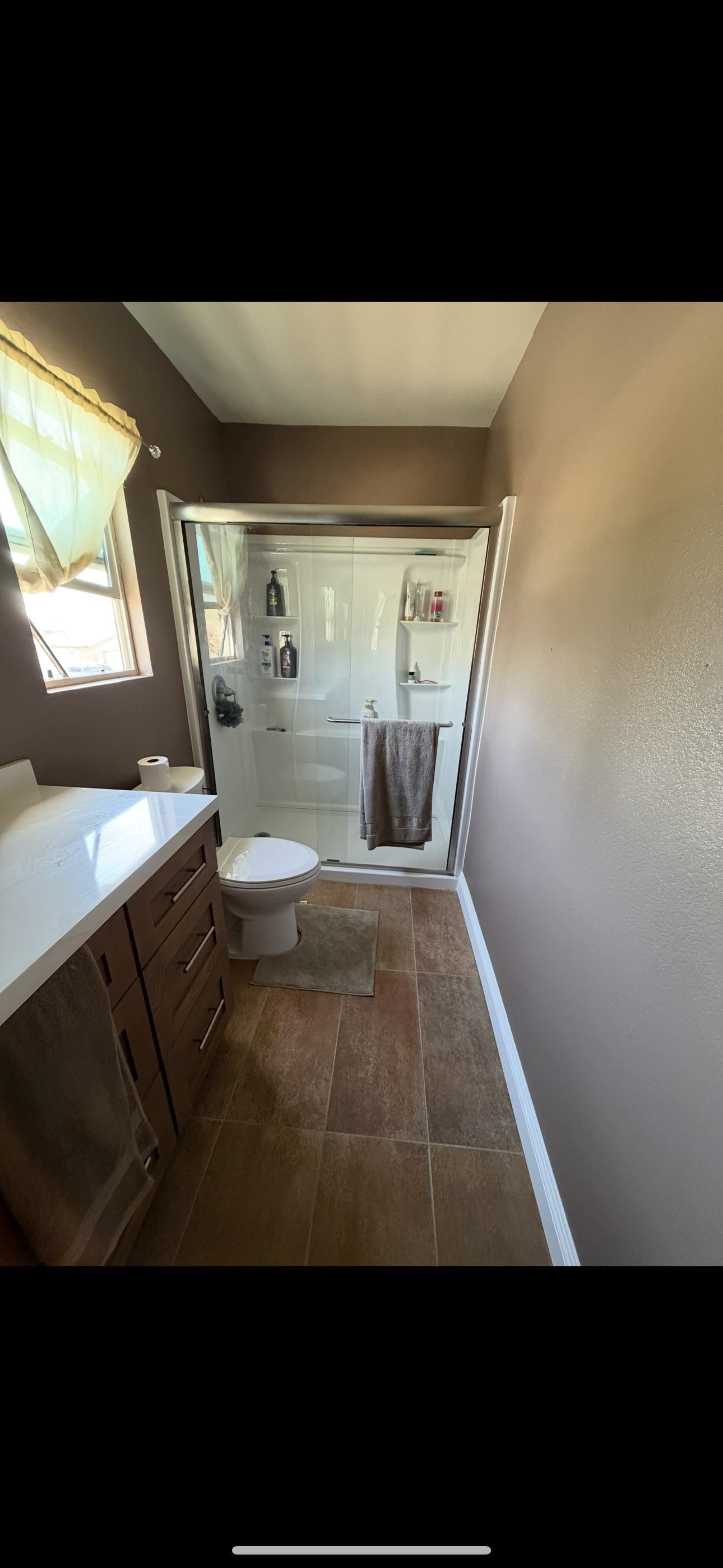 Bathroom with brown walls, window with light green curtain, toilet, beige tiled floor, wooden vanity with a white countertop, and a glass shower with shelves and towels.