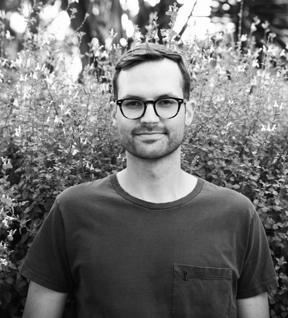 Black and white photograph of a young man with glasses and a beard, smiling and standing in front of bushes.