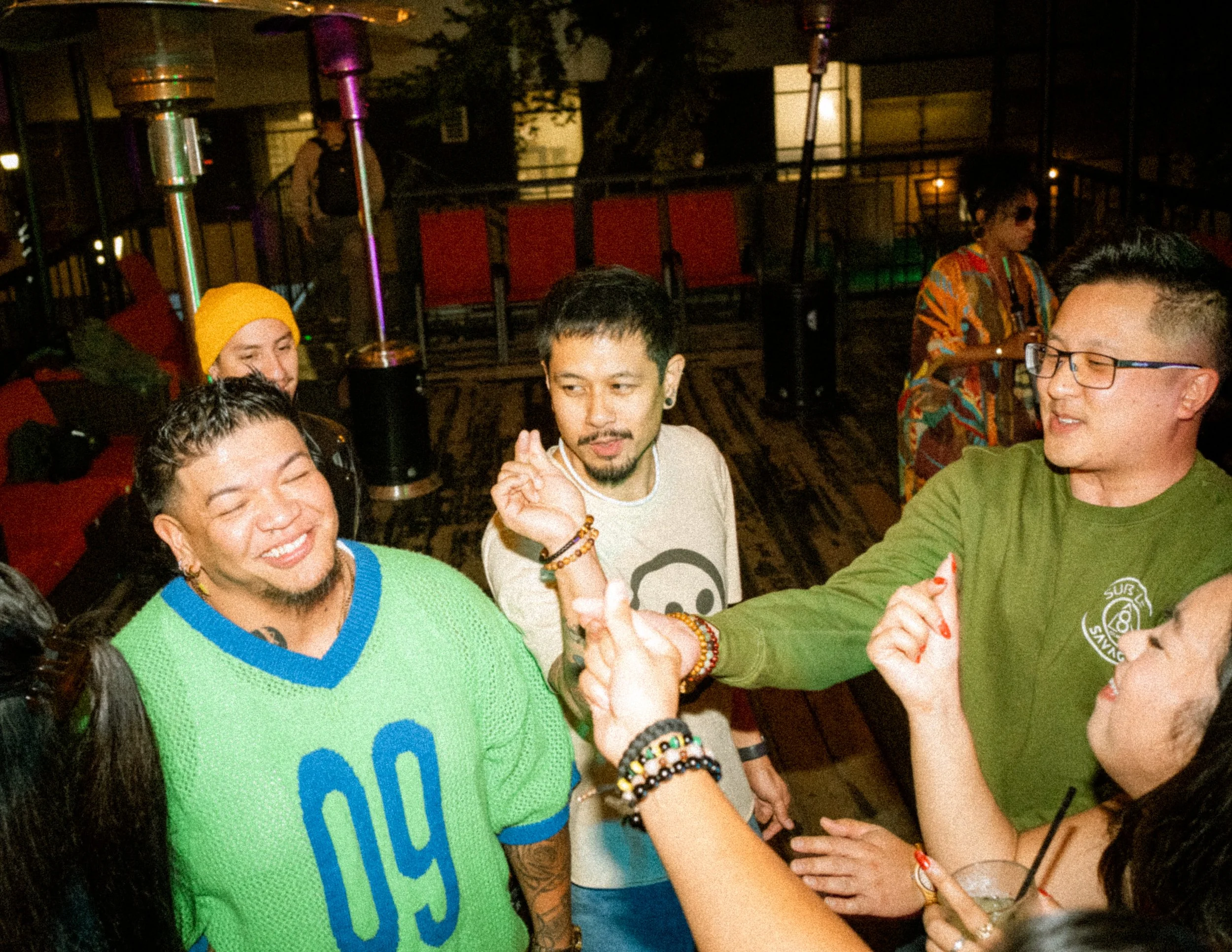 Group of people socializing at a gathering, some wearing colorful clothing, in a dimly lit indoor space.