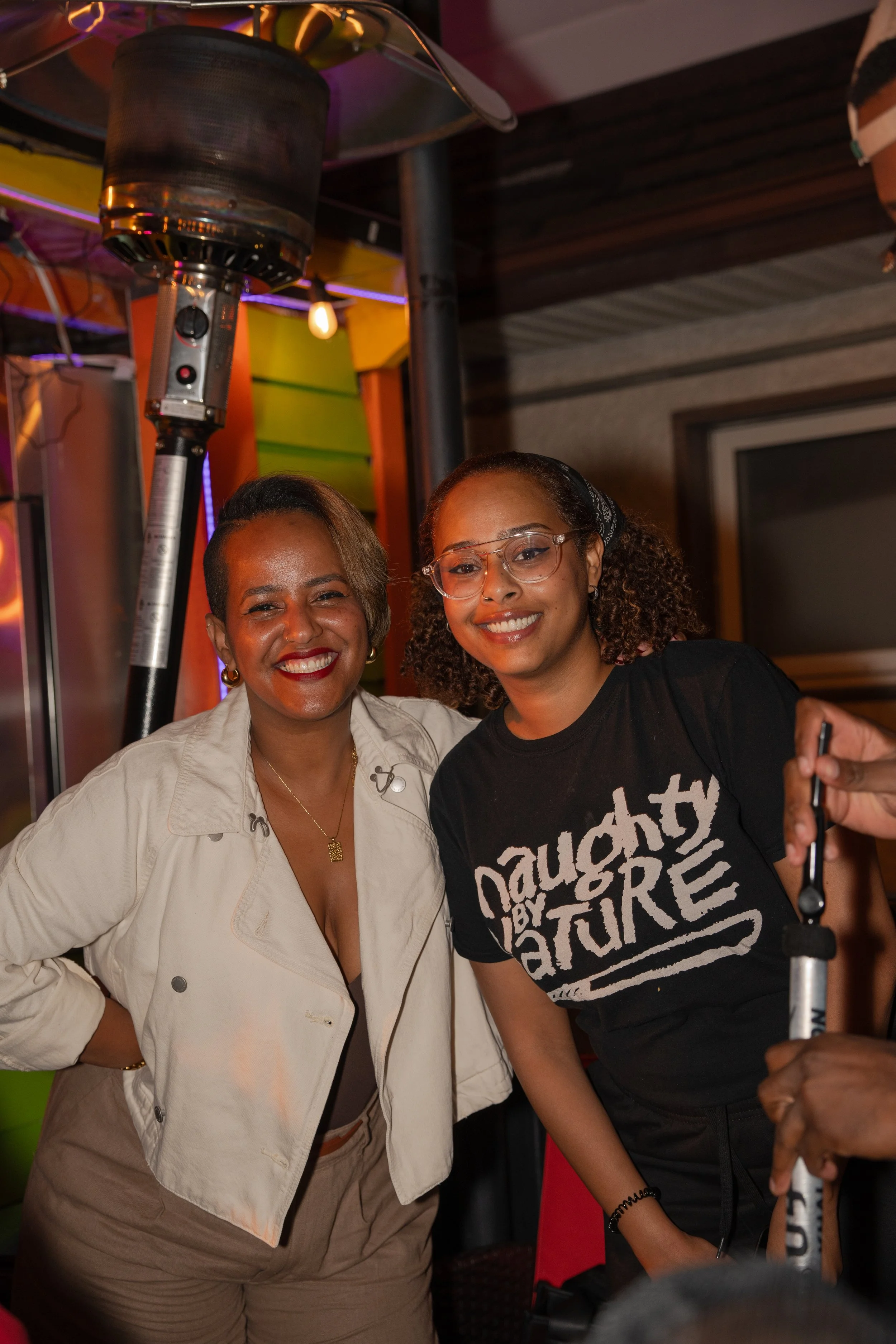 Two women smiling and posing for a photo at an indoor gathering or party, with a patio heater and colorful background behind them.