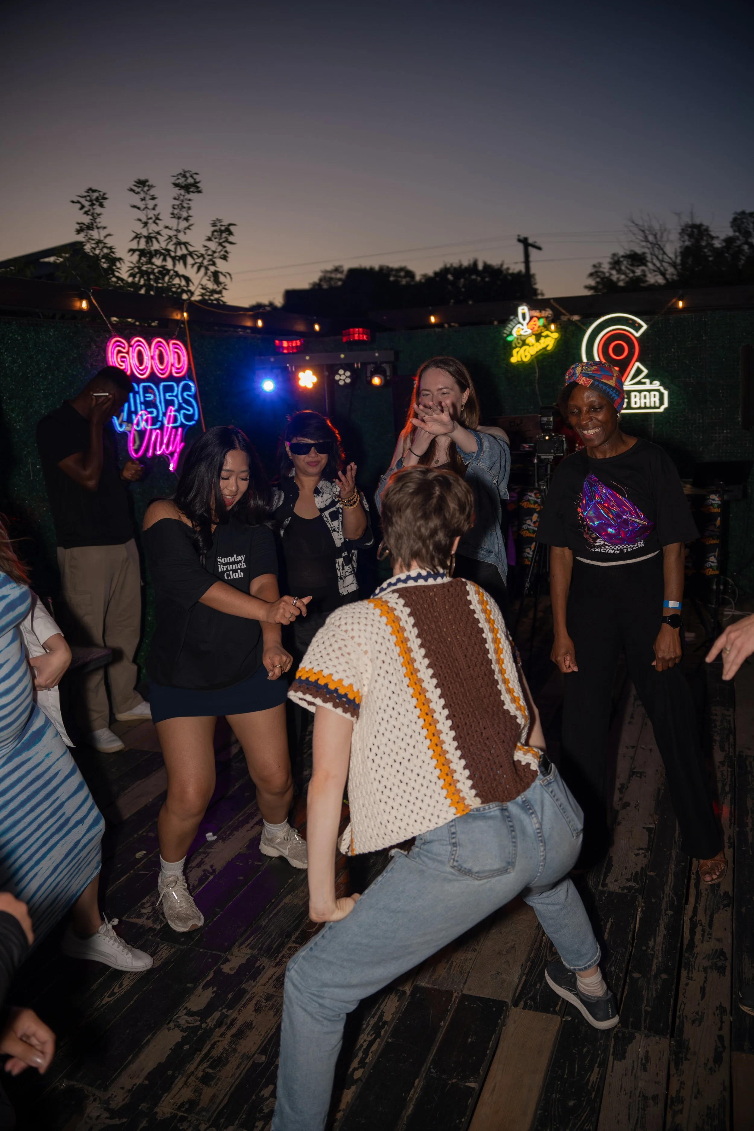 People dancing and having fun at a nighttime outdoor bar with neon signs.