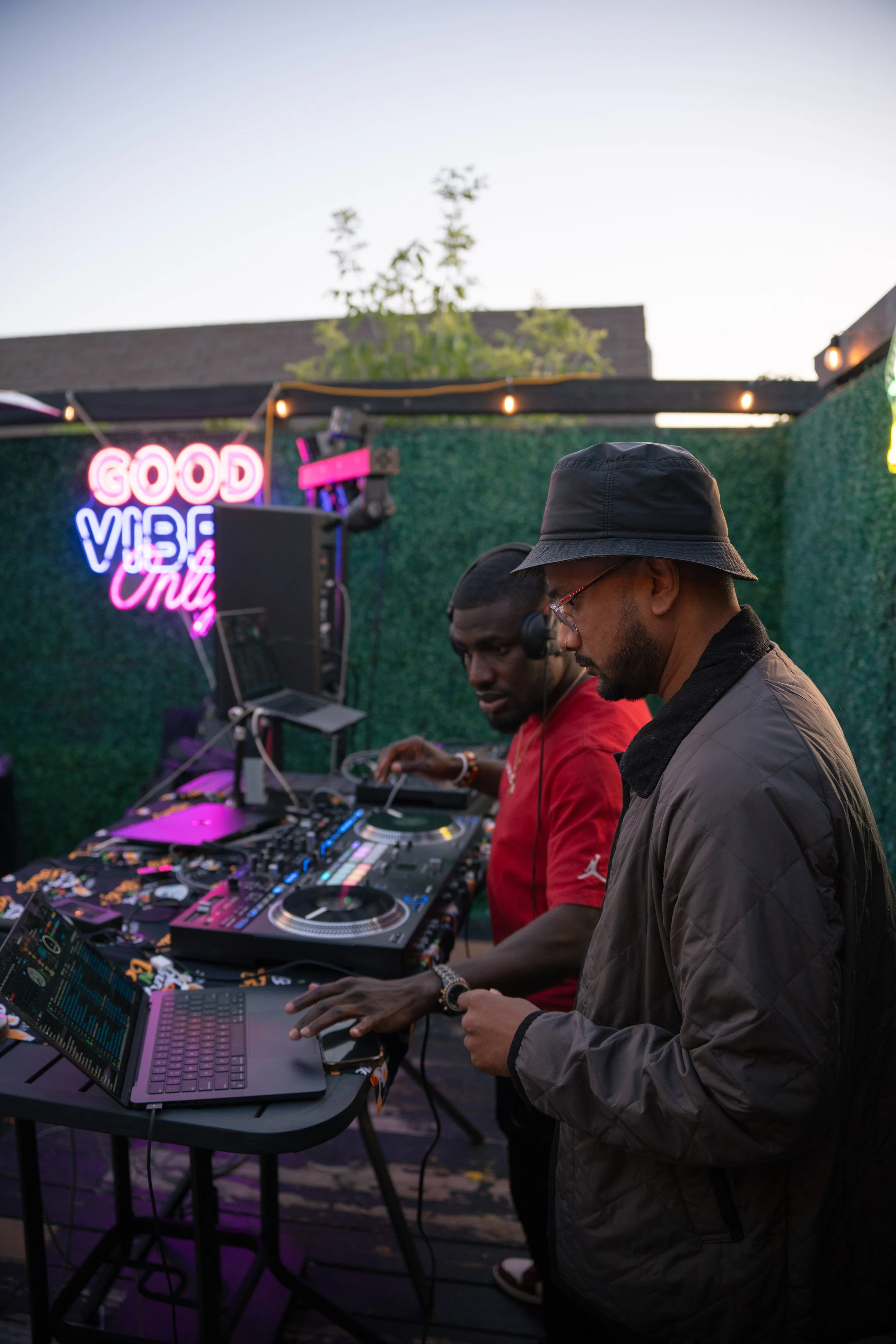 Two DJs performing at an outdoor event during the evening, with a neon sign that says 'Good Vibe Only' in the background.