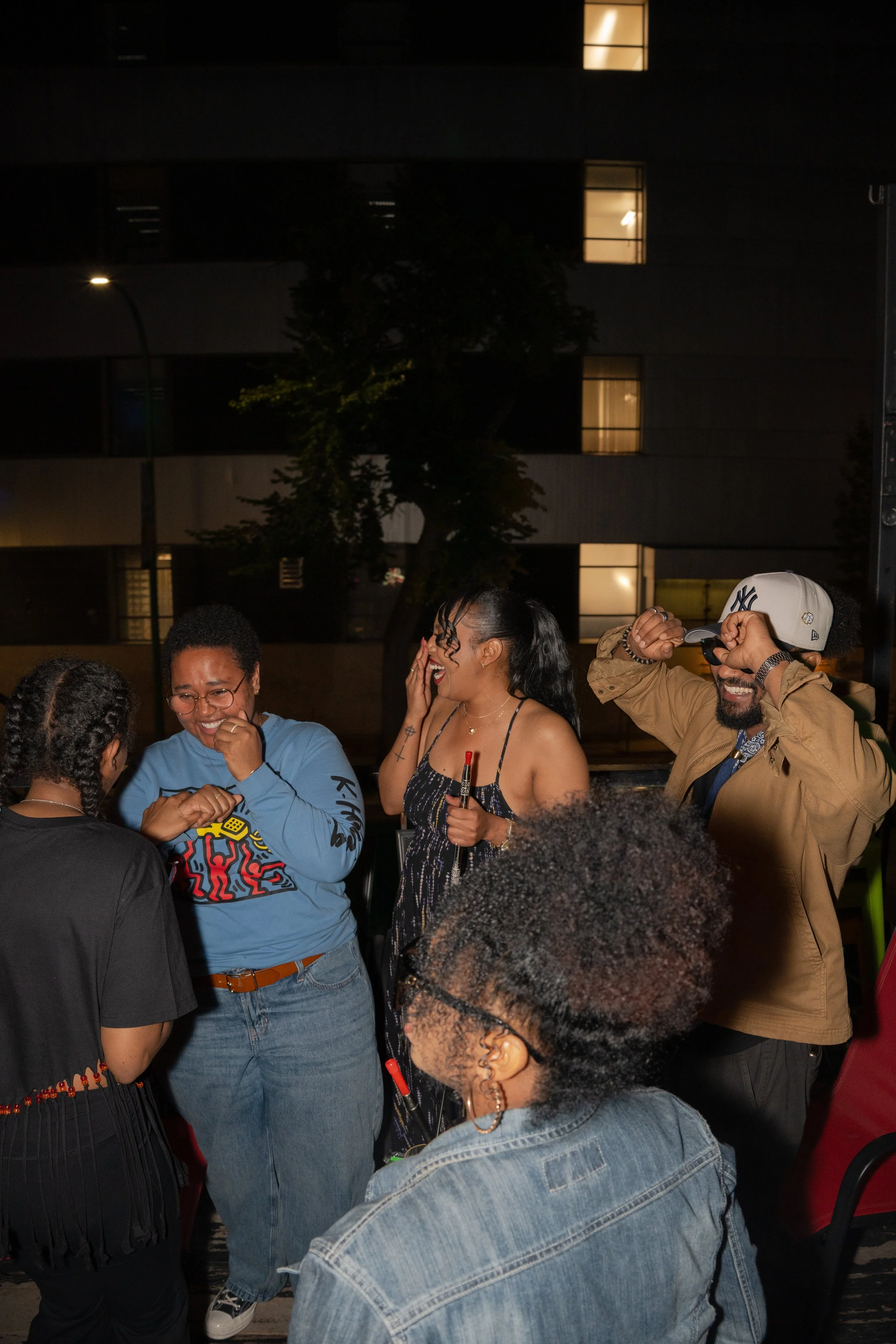 Group of friends laughing and talking at an outdoor night gathering, with a building in the background.