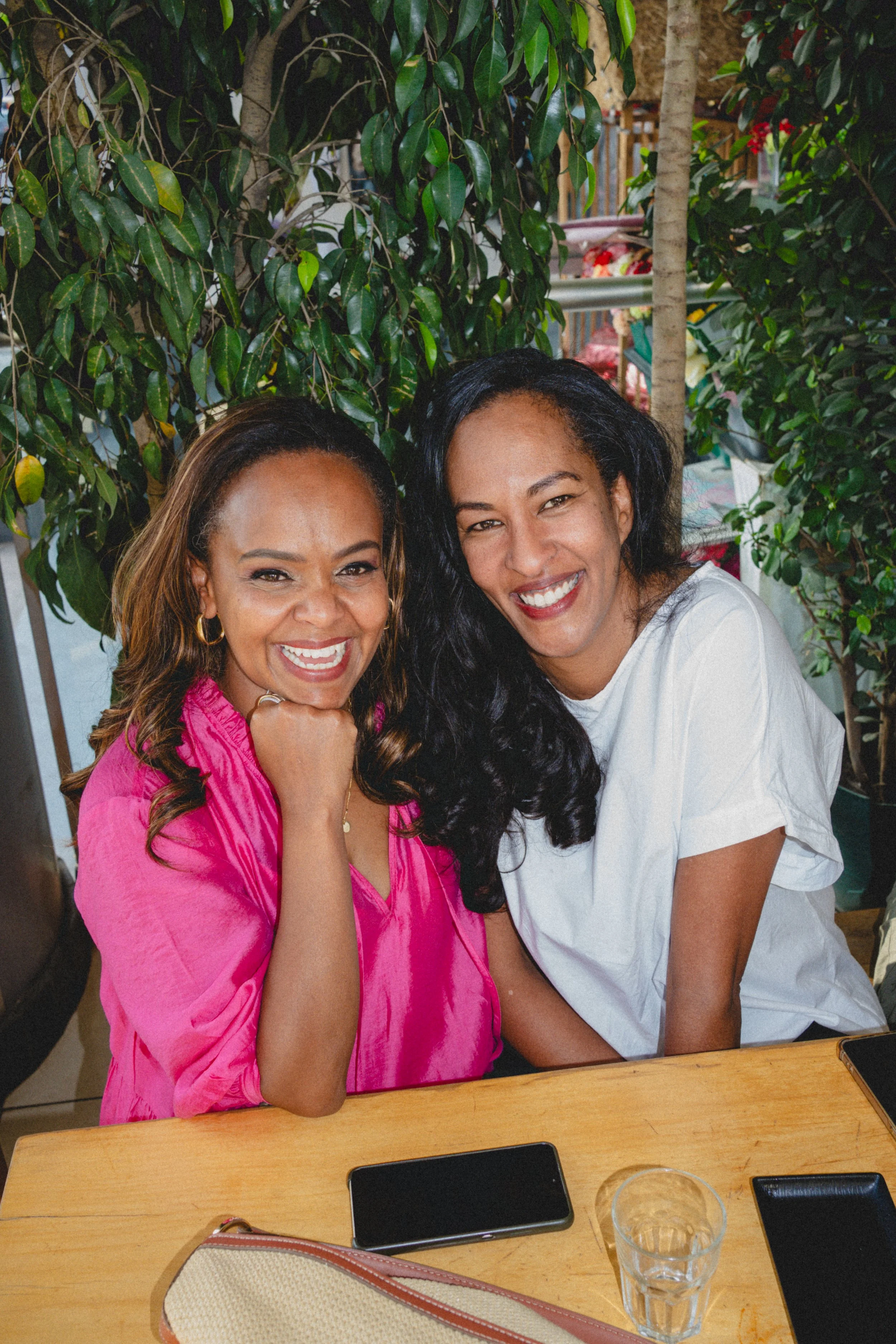 Two women sitting at a wooden table with greenery in the background, smiling at the camera.