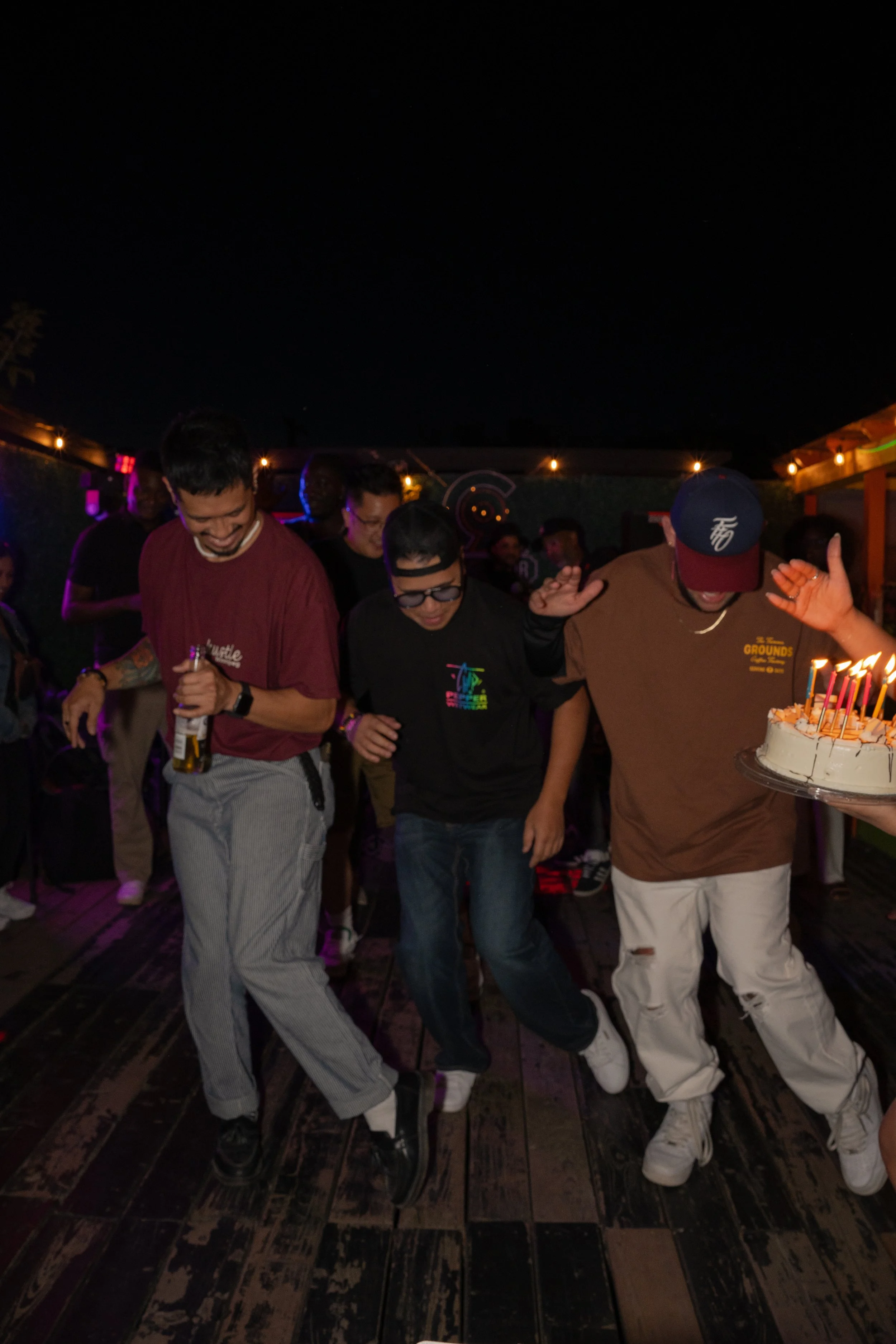 Three men dancing at a birthday party at night, with a birthday cake with lit candles held by one man.