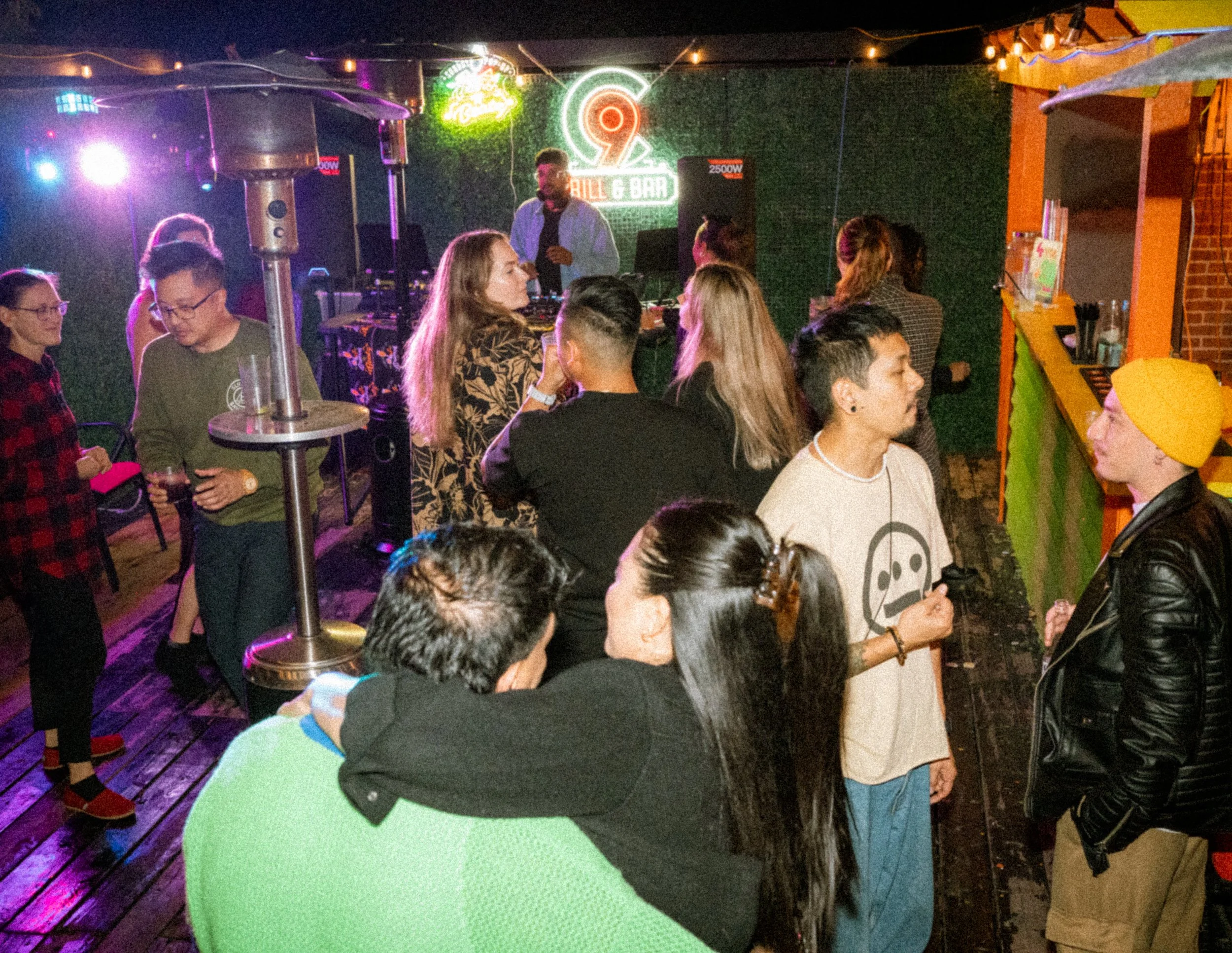 People socializing at a nightclub with a DJ playing at the back, neon signs, and colorful lighting.