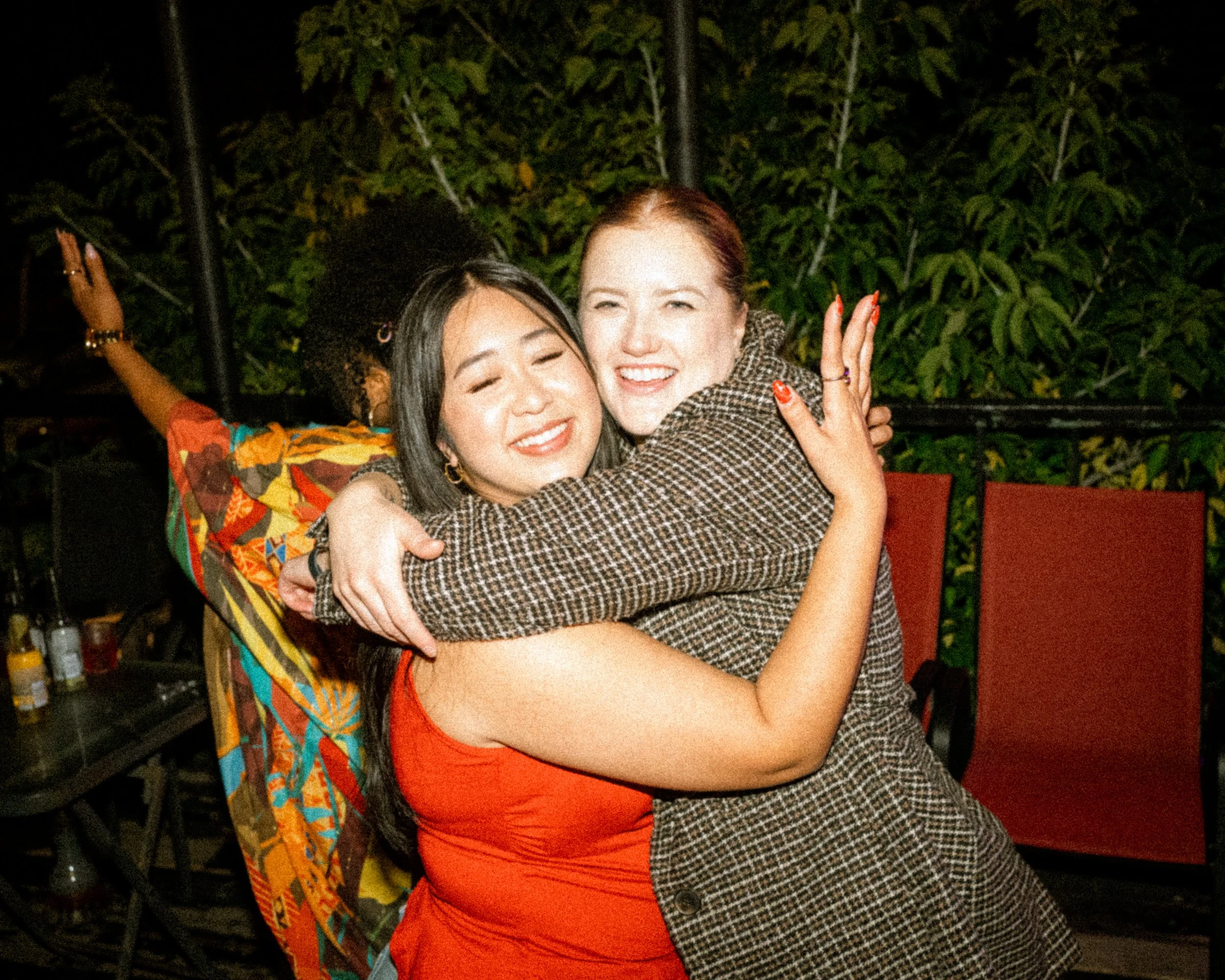 Two women hugging and smiling at night indoors, with plants and chairs in the background.