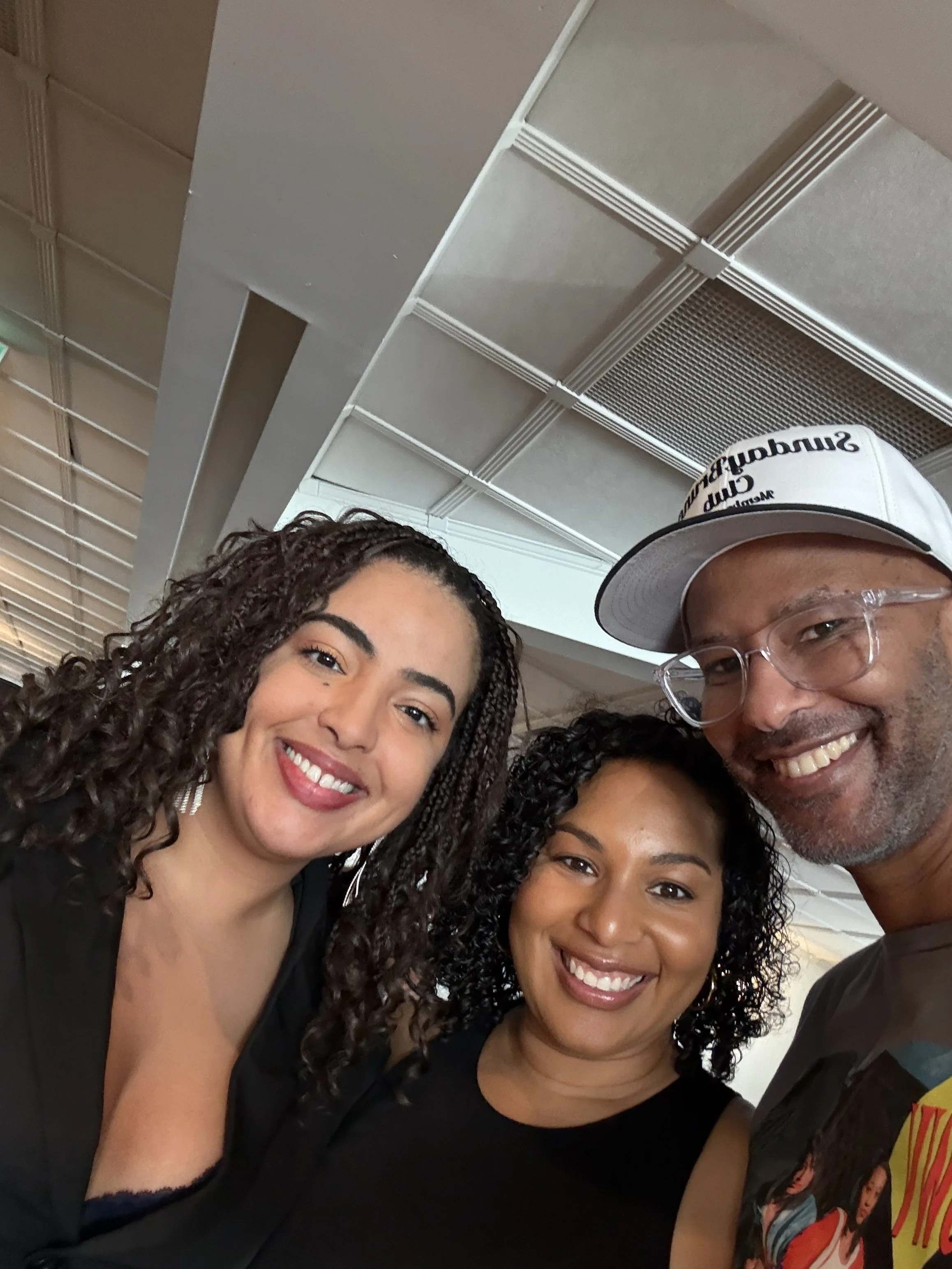 Three smiling people posing for a photo indoors. Two women and one man, all with curly hair, dressed casually, smiling at the camera.