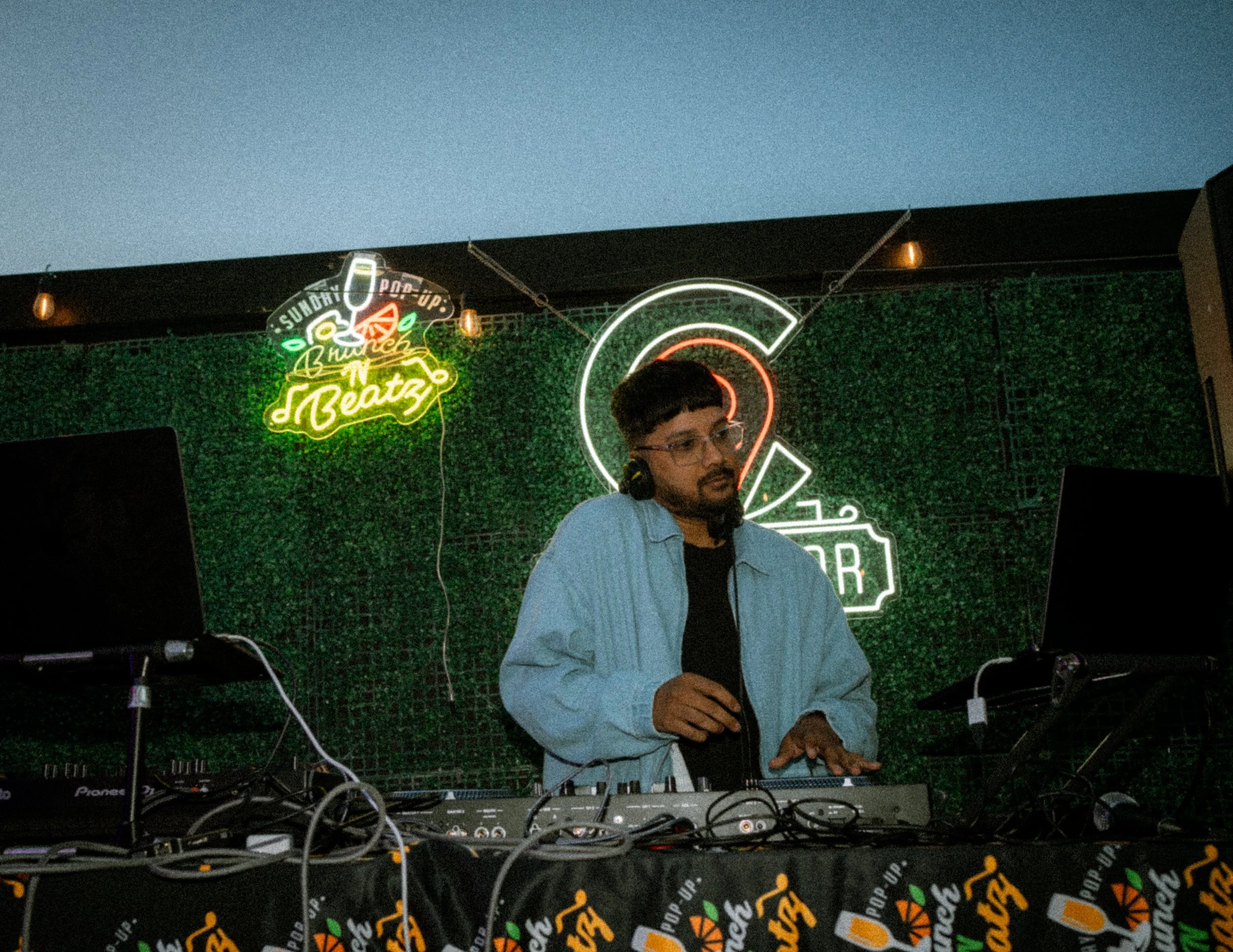 A DJ with glasses and dark hair, wearing a blue jacket, stands behind a DJ setup with turntables and laptops. Neon signs in the background include a tropical bird and the words 'Sunday pop-up' and 'Brunch & Beatz' with a green hedge wall.