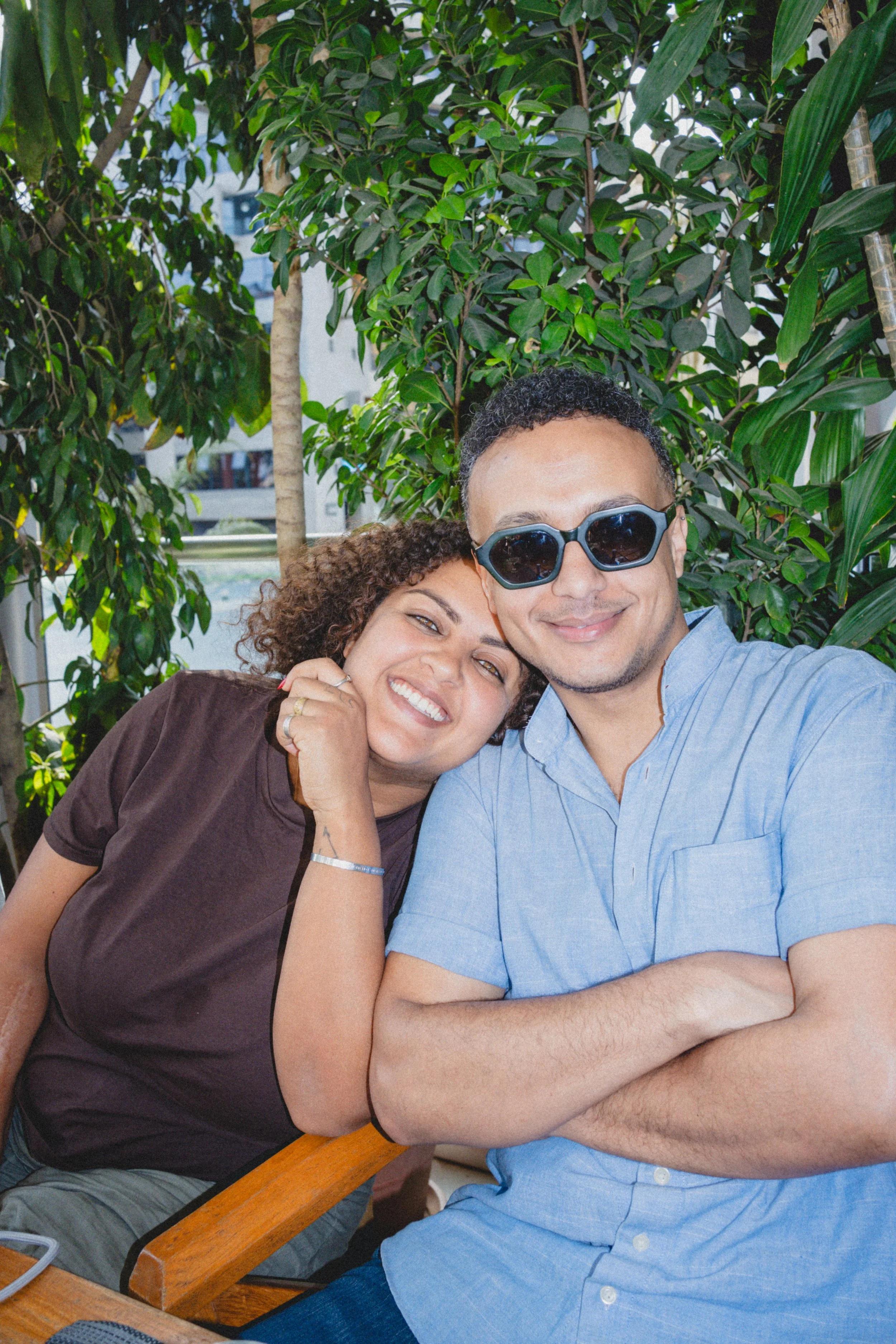 A man and woman sitting close together outdoors, smiling, with greenery in the background.