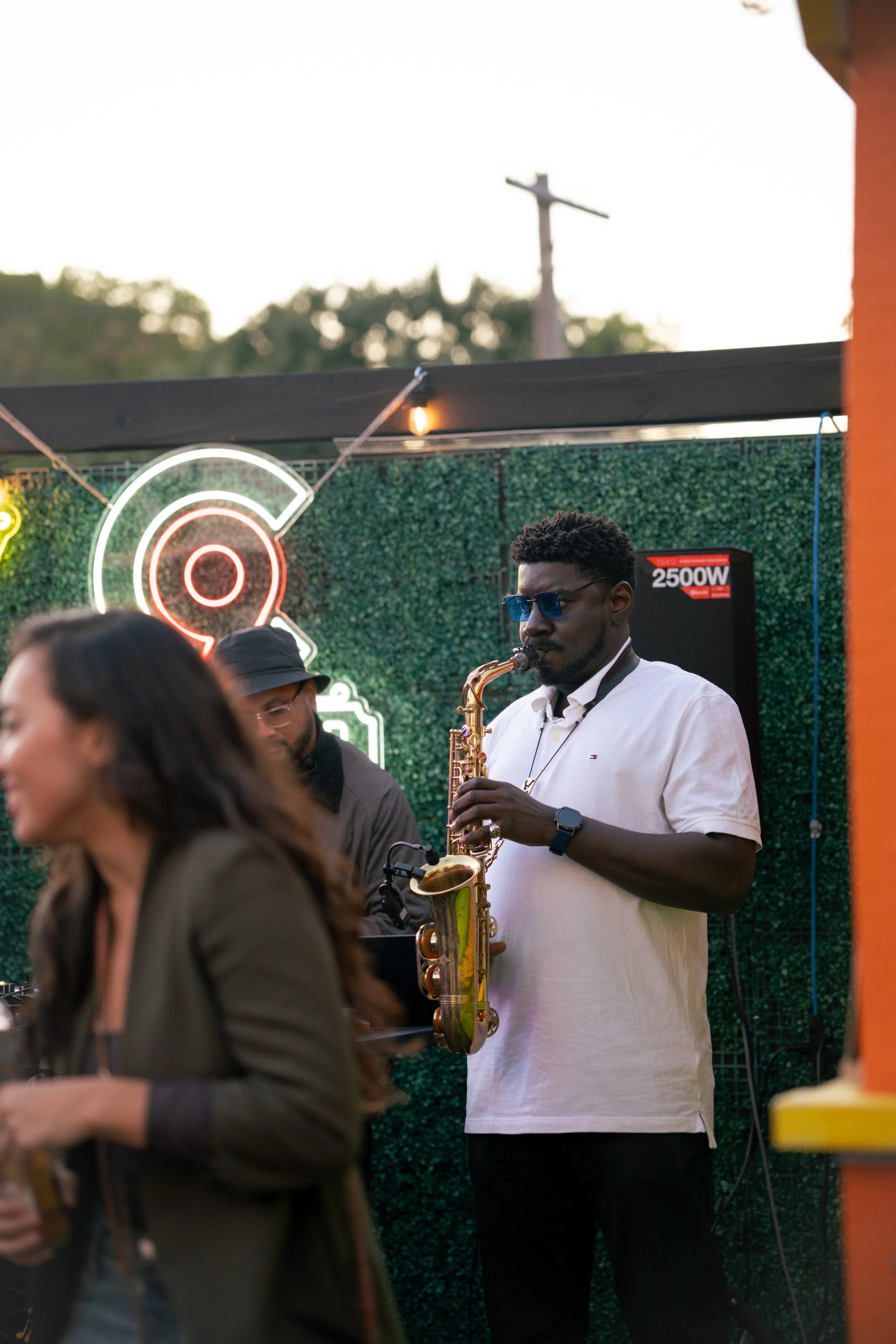 A man playing a saxophone at an outdoor event during the evening. A woman is in the foreground, out of focus. There is a green backdrop with neon signs and a speaker in the background.