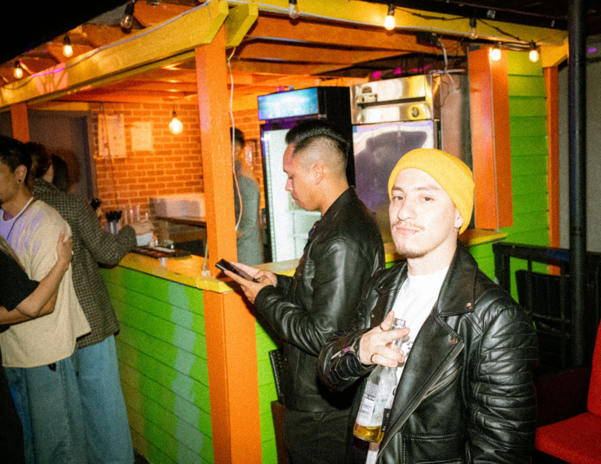 Two young men standing at a bar counter in a colorful, lively venue. One is wearing a yellow beanie and a black leather jacket, holding a drink and making a peace sign. The other is wearing a leather jacket, looking at his phone.