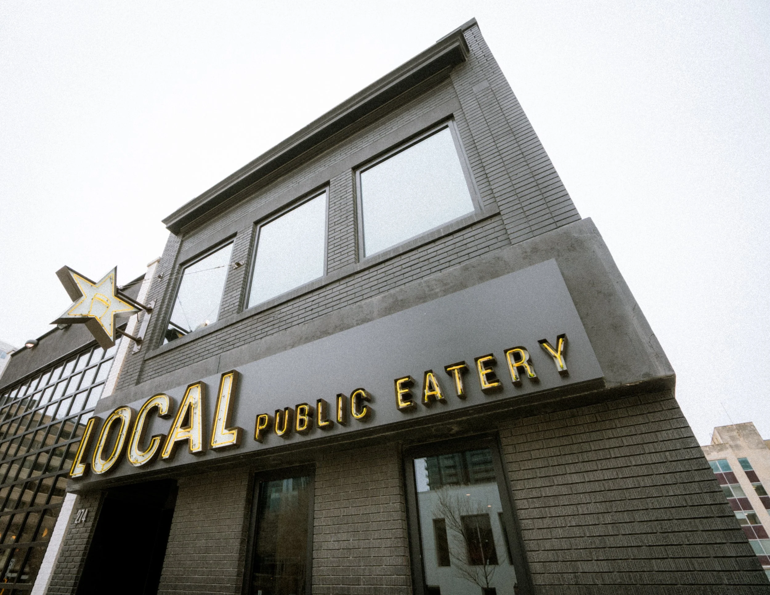 Exterior of a building with a sign that reads 'LOCAL PUBLIC EATERY' in yellow letters, with a star-shaped lit sign on the side.
