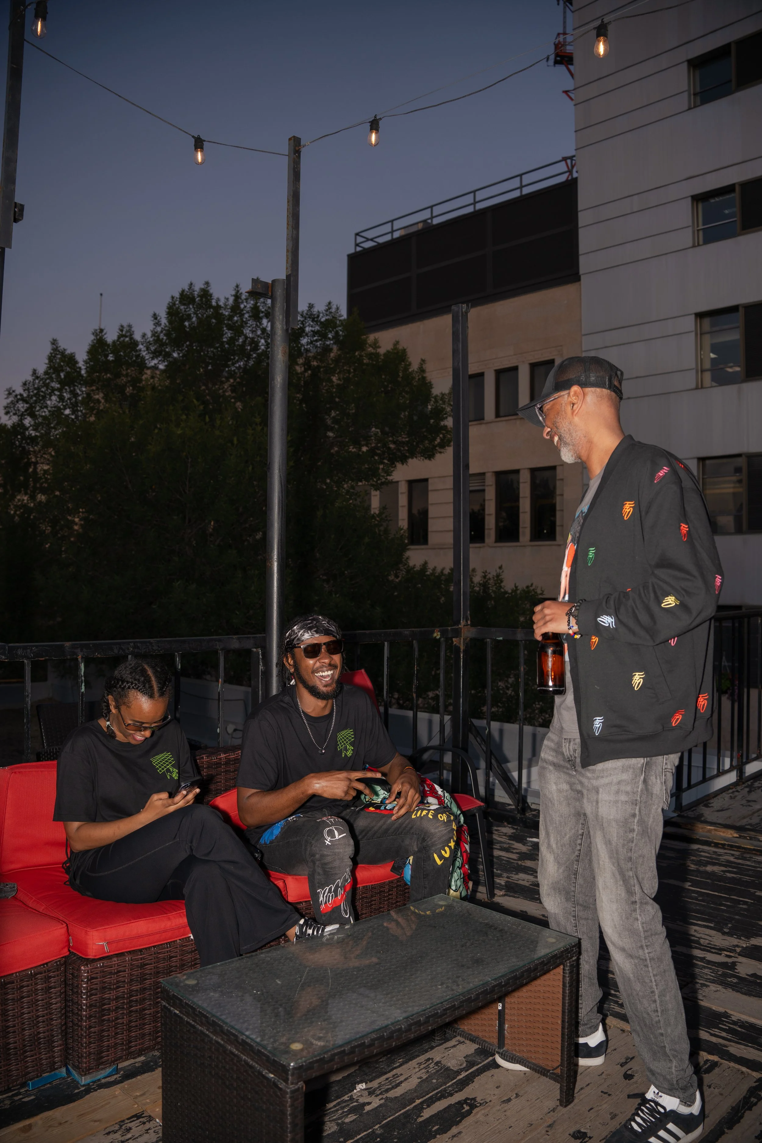 Three friends socializing on a rooftop terrace at dusk, with string lights overhead, sitting on a wicker couch with red cushions, laughing and chatting, with a glass table in front of them and a city building in the background.