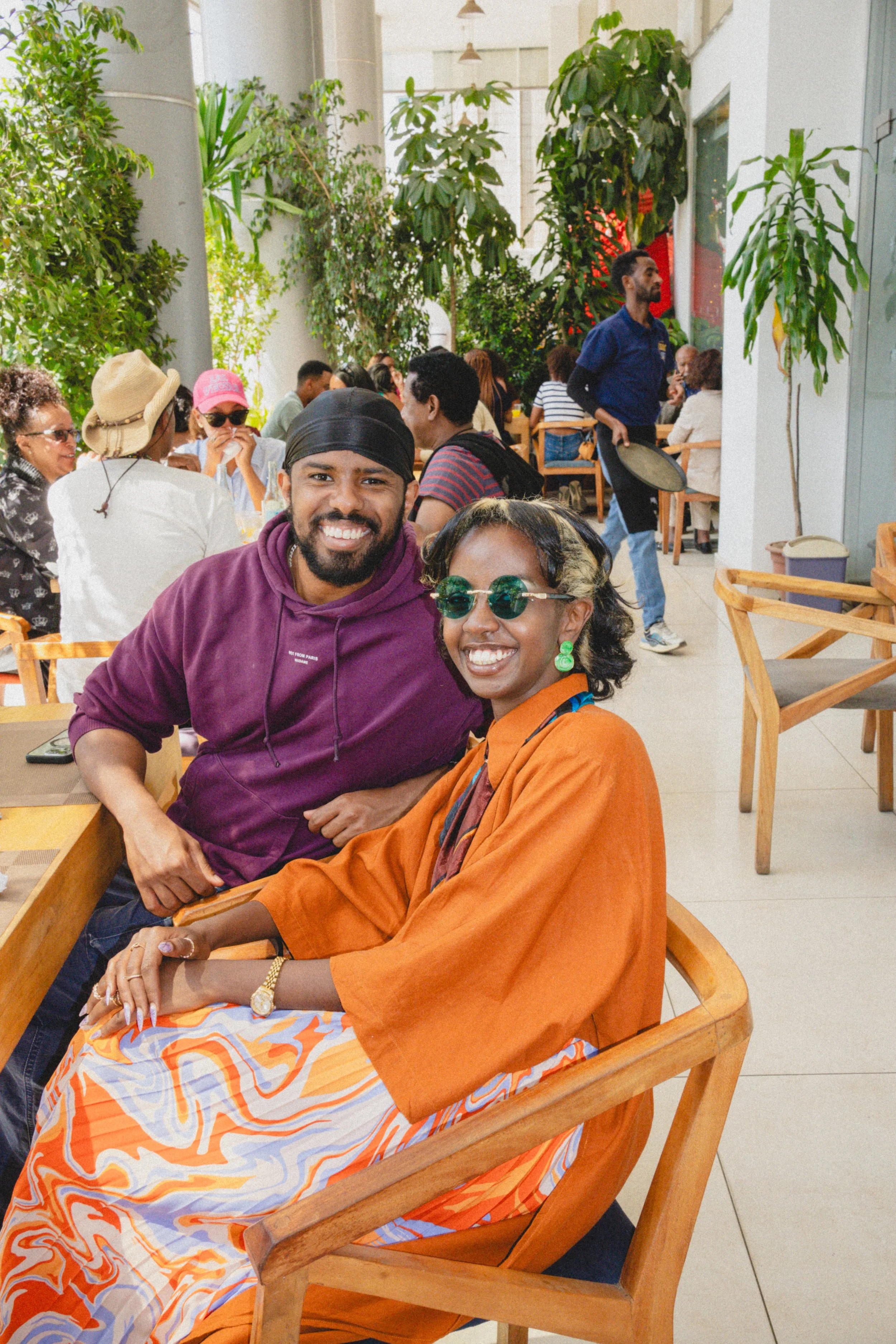 Two people sit together and smile in a busy indoor restaurant with green plants and people dining in the background.