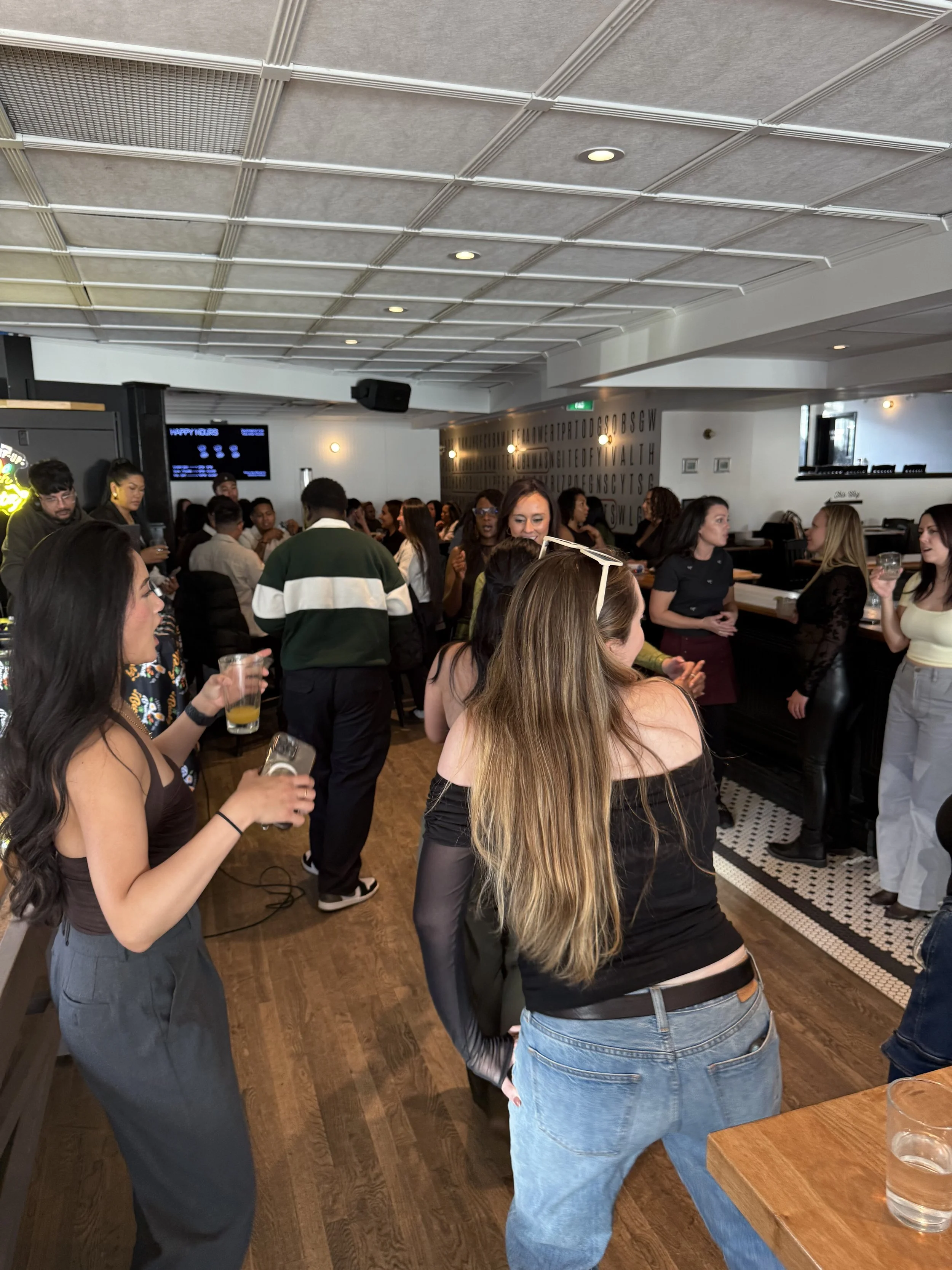 People socializing at a lively indoor gathering, some holding drinks, in a restaurant or bar with modern decor.