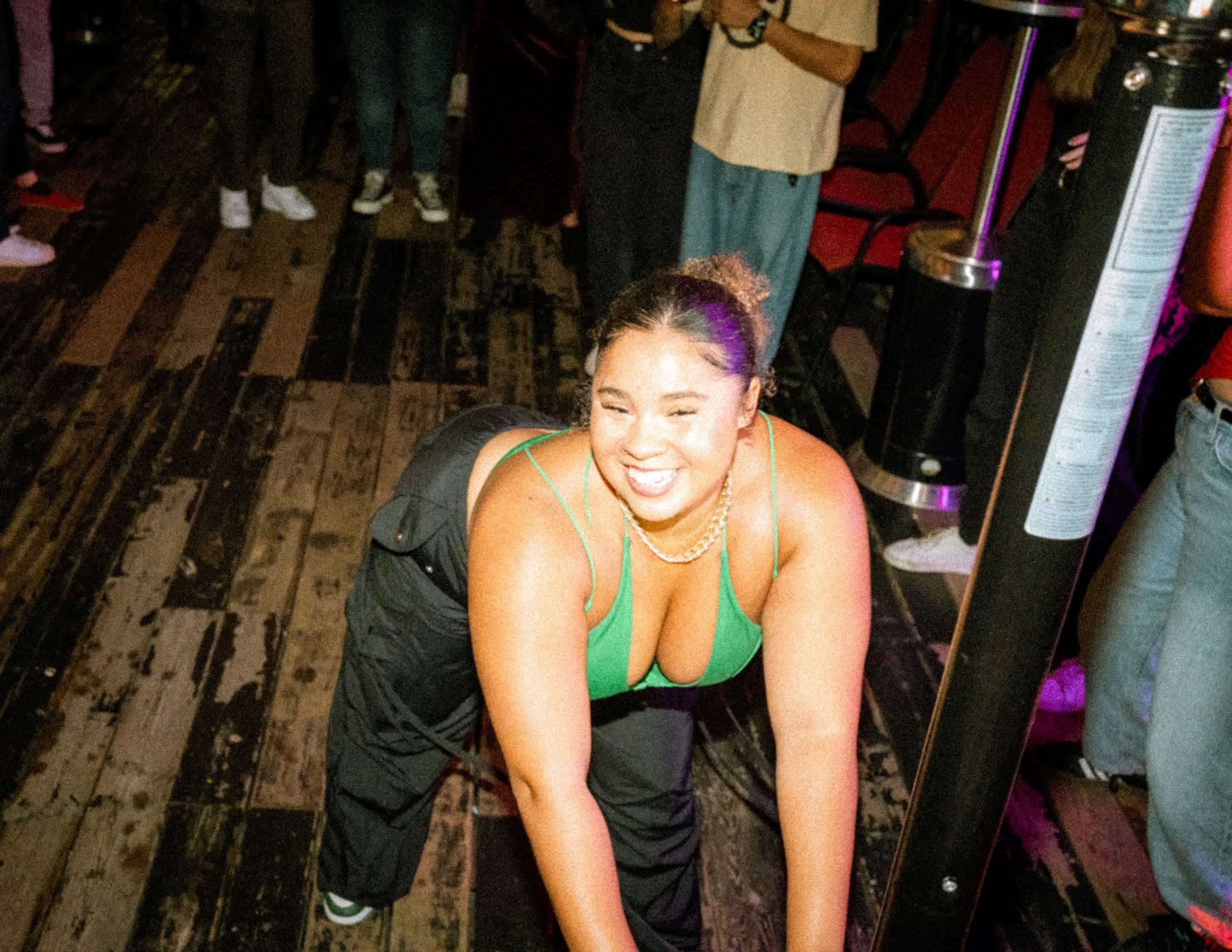 A woman with curly hair in a topknot, smiling, and wearing a green tank top and black pants, crouching on a wooden floor at a social event with people around.