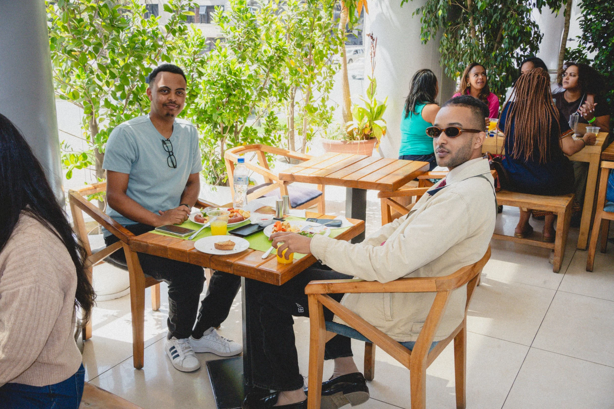 A group of people dining at a restaurant with green plants in the background. Two men are sitting at a table in the foreground, one with sunglasses and the other with glasses hanging from his shirt. There are plates of food, drinks, and utensils on t