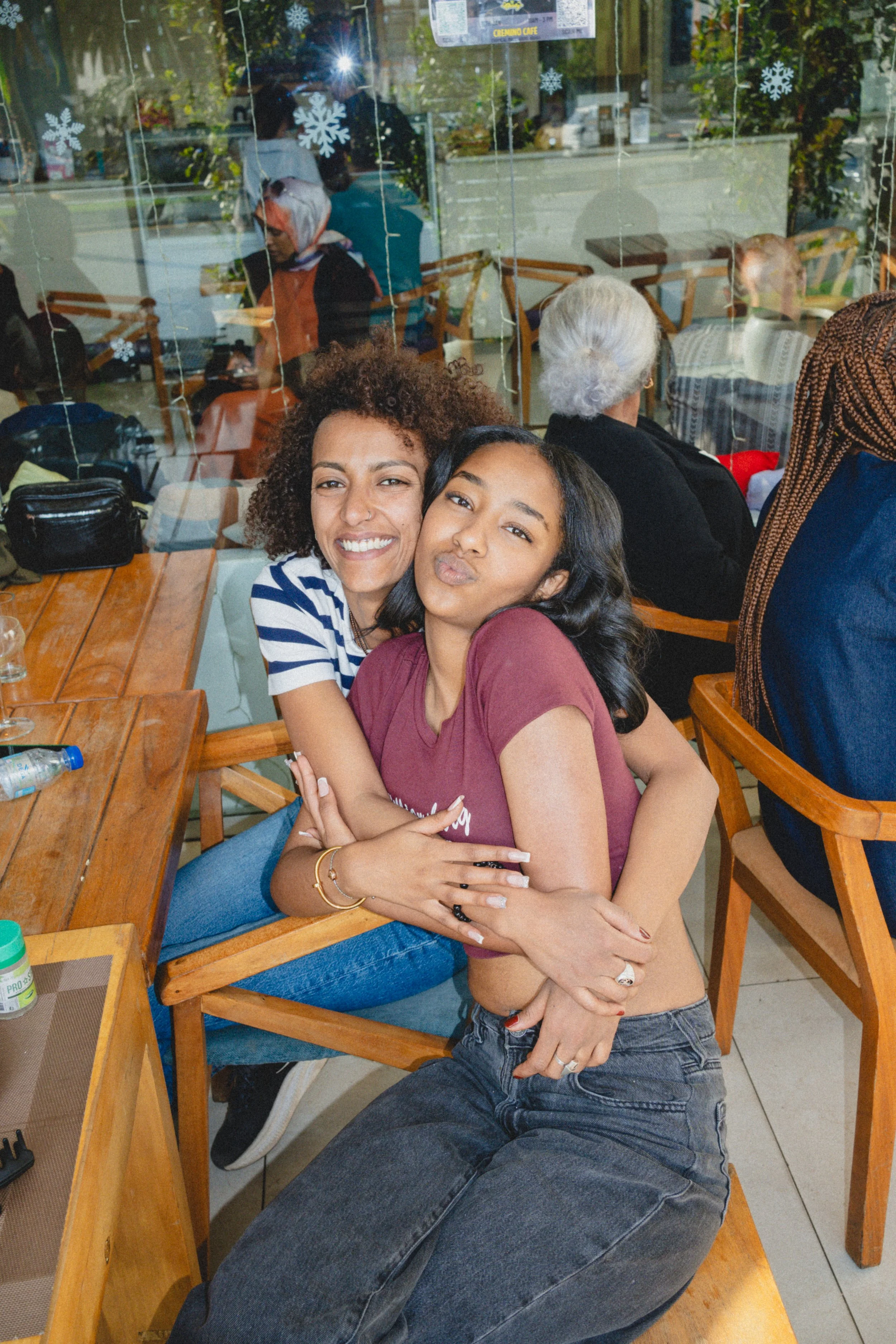 Two young women hugging and smiling inside a cafe, with other people seated at tables and a glass window behind them reflecting the street.