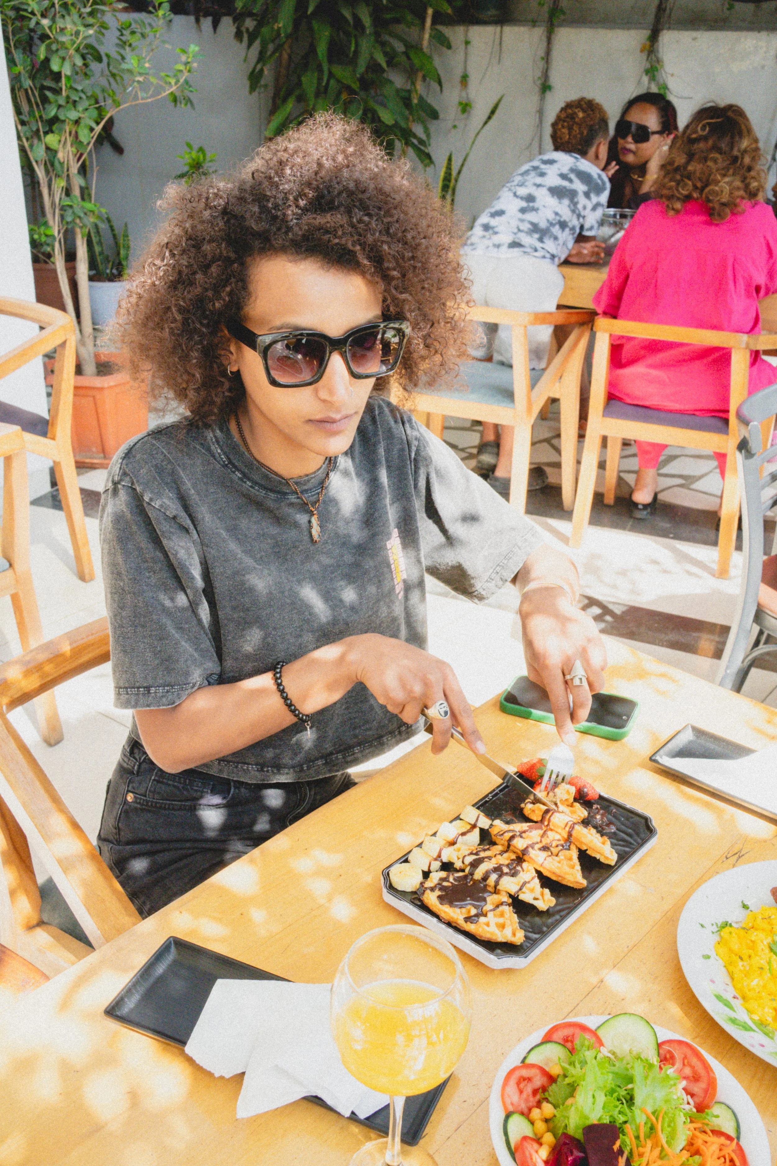 Woman with curly hair and large sunglasses eats waffles with chocolate syrup at an outdoor restaurant table, with a salad and drink nearby.