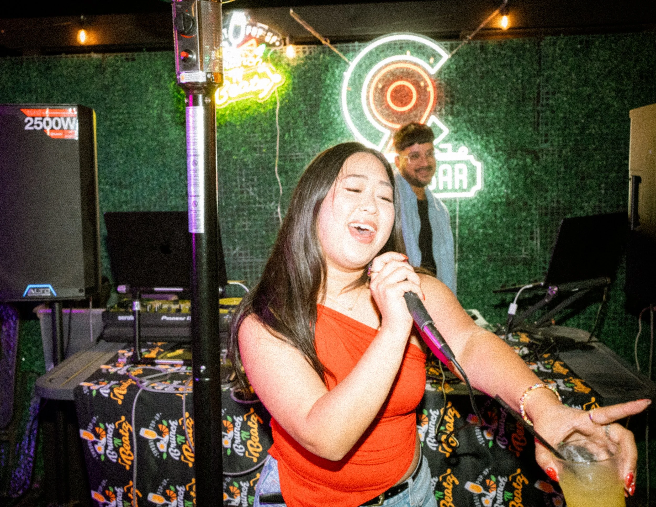 A woman singing into a microphone at a party or nightclub with a DJ in the background, neon signs, and a green textured wall.