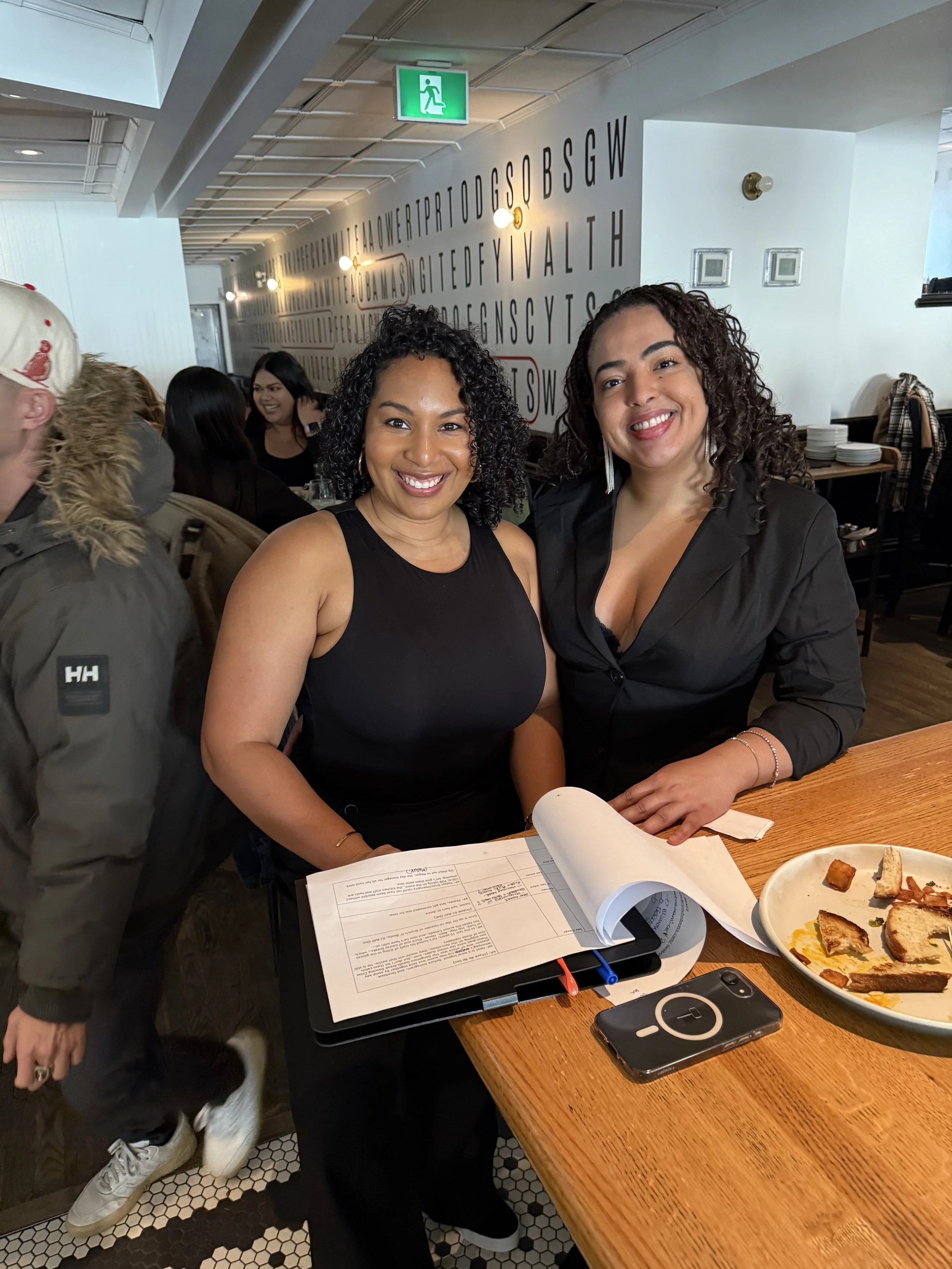 Two women smiling at a restaurant or cafe table, with papers, a phone, and plates of food in front of them, with a patterned floor and a wall decorated with alphabet letters in the background.