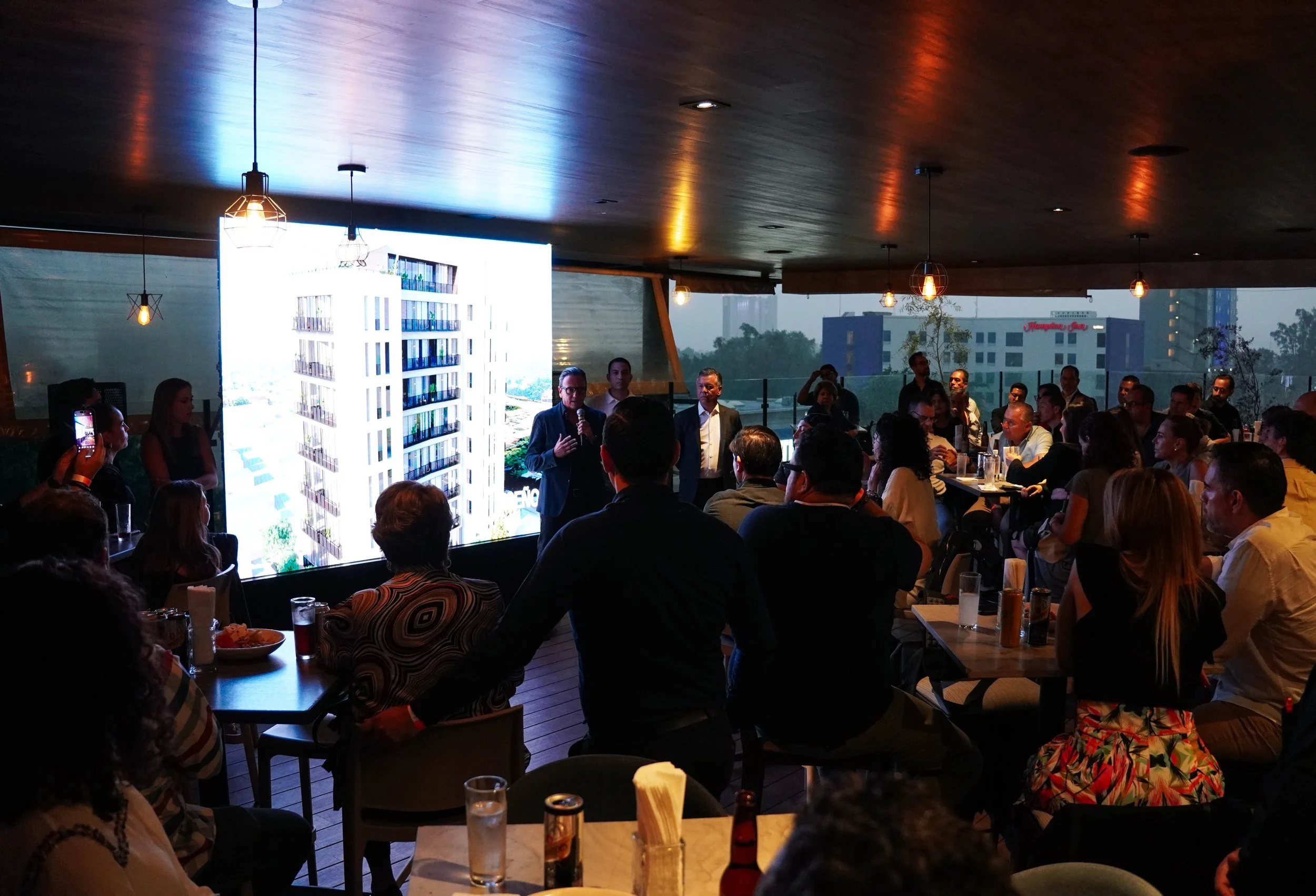 Imagen de una reunión o presentación en un restaurante o salón de eventos con muchas personas sentadas, frente a un orador que está de pie frente a una pantalla grande con una imagen de un edificio de apartamentos.