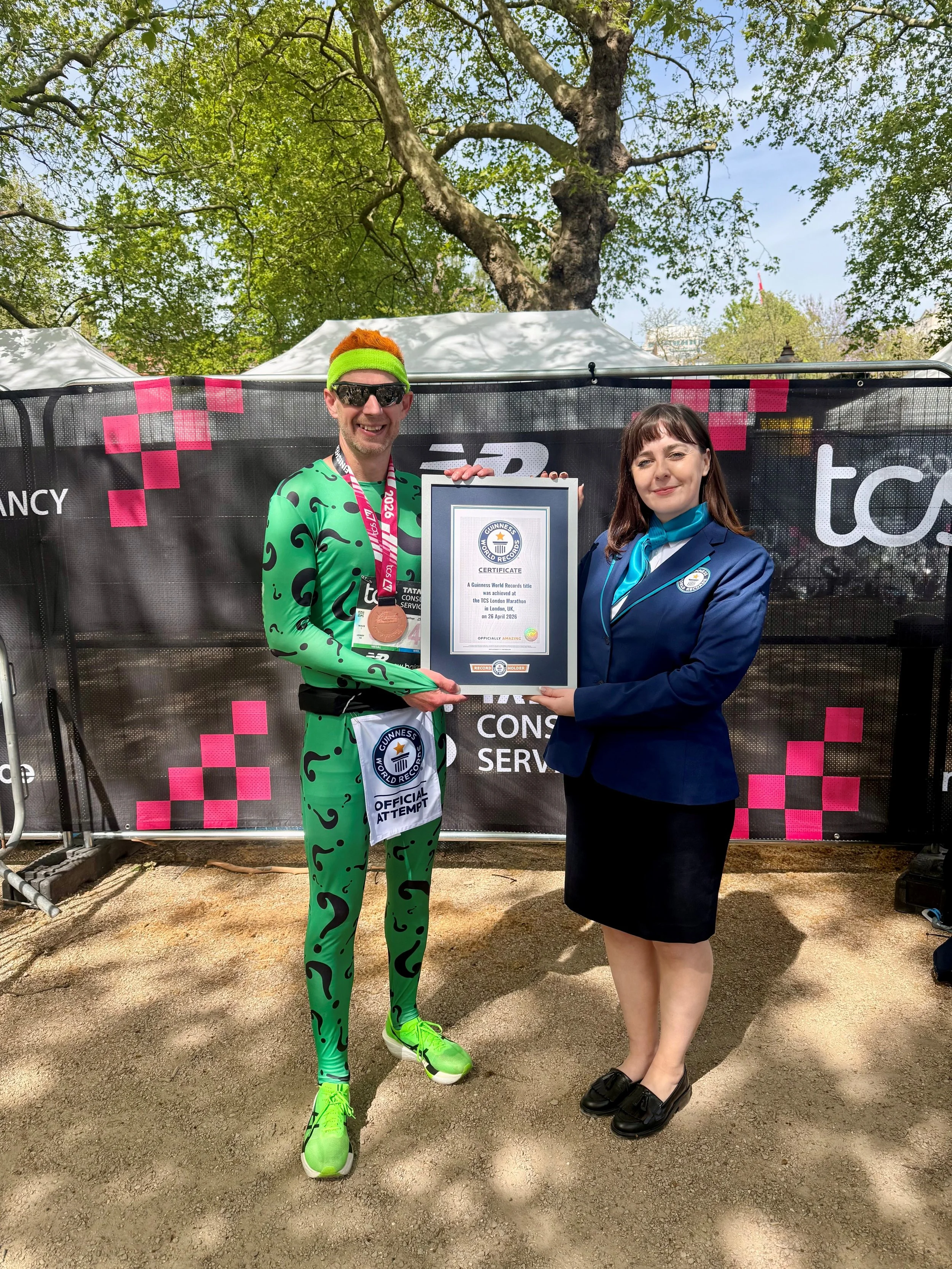 A man in a green jog suit with black question marks, holding a Guinness World Record certificate and medal, standing next to a woman in a navy blue blazer and skirt, outdoors in front of a black and pink checkered banner, with trees overhead.