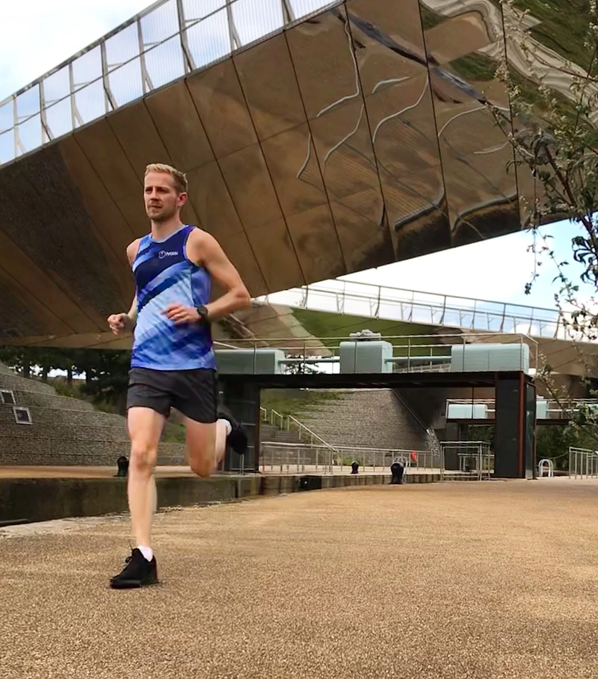 A male athlete running on a dirt track in a sports stadium, with large reflective building structure in the background and some green foliage.