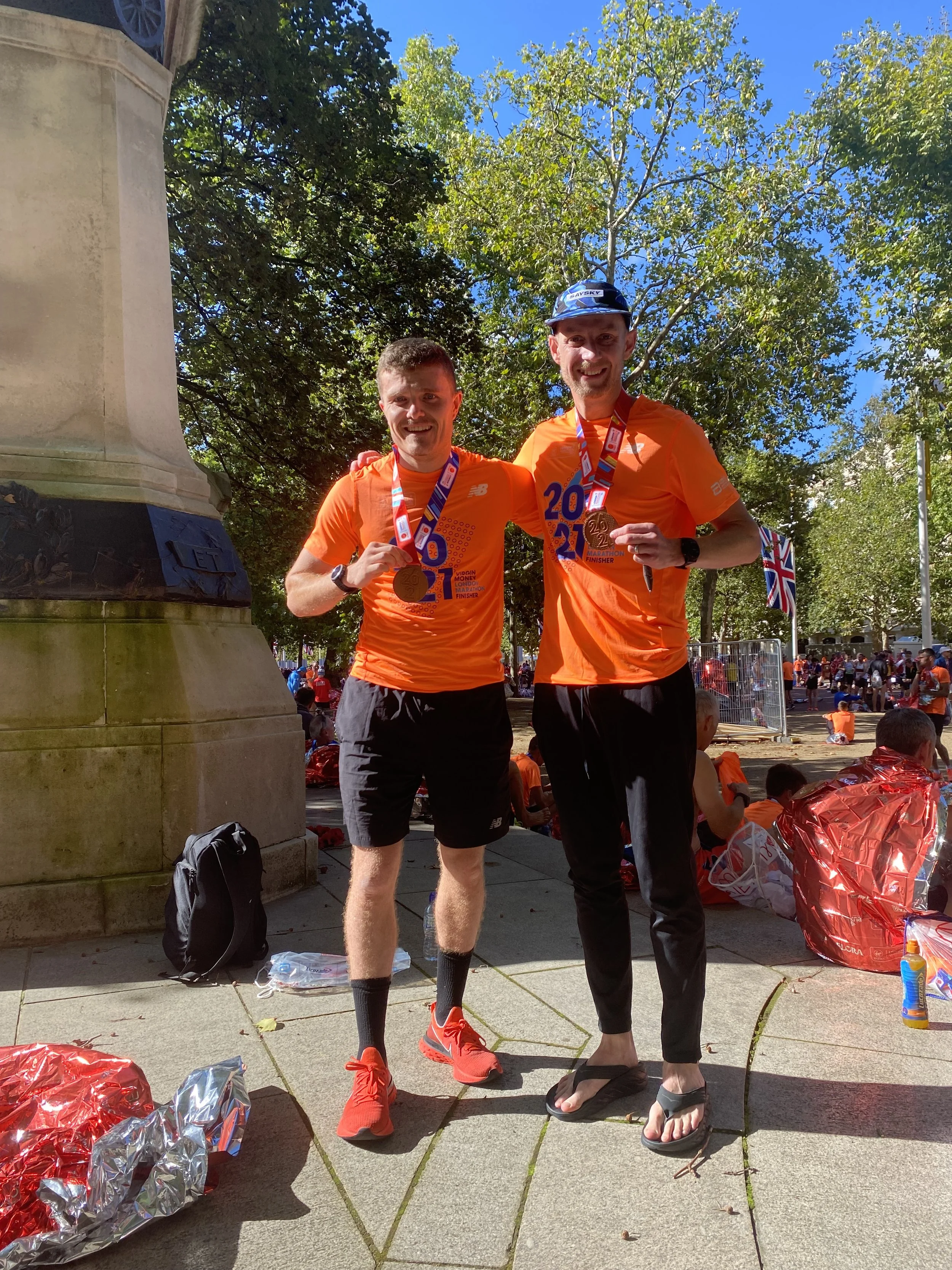 Two men in orange athletic shirts holding medals and smiling, celebrating a race finish, standing outdoors near a stone monument with trees and a crowd in the background.