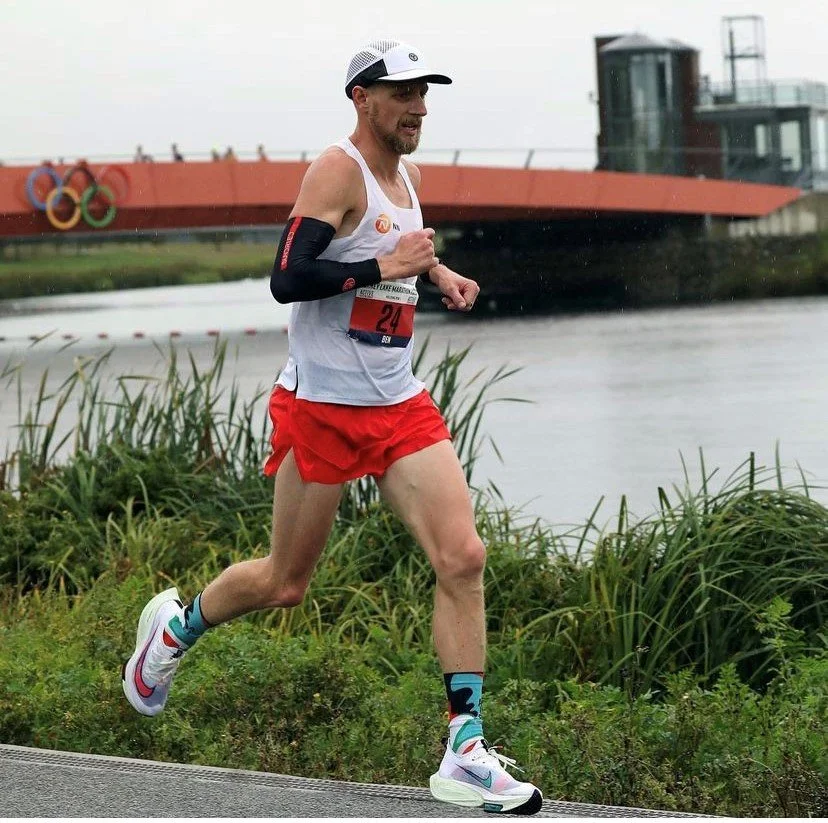 A male runner in athletic gear running outdoors near water with grasses in the foreground and a bridge with Olympic rings in the background.