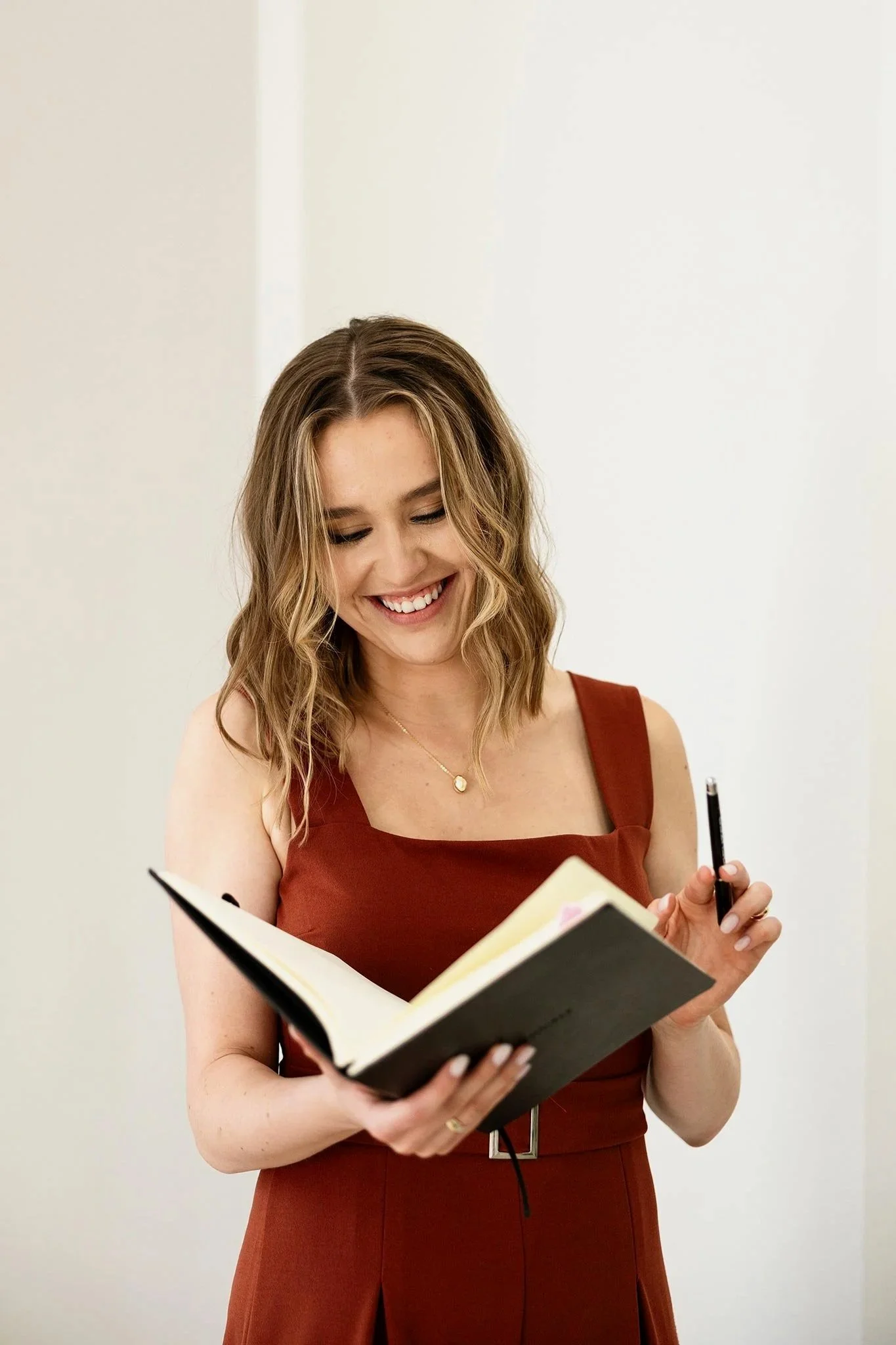 A woman with shoulder-length wavy hair wearing a sleeveless rust-colored dress, smiling as she looks at her open notebook while holding a pen in her right hand.