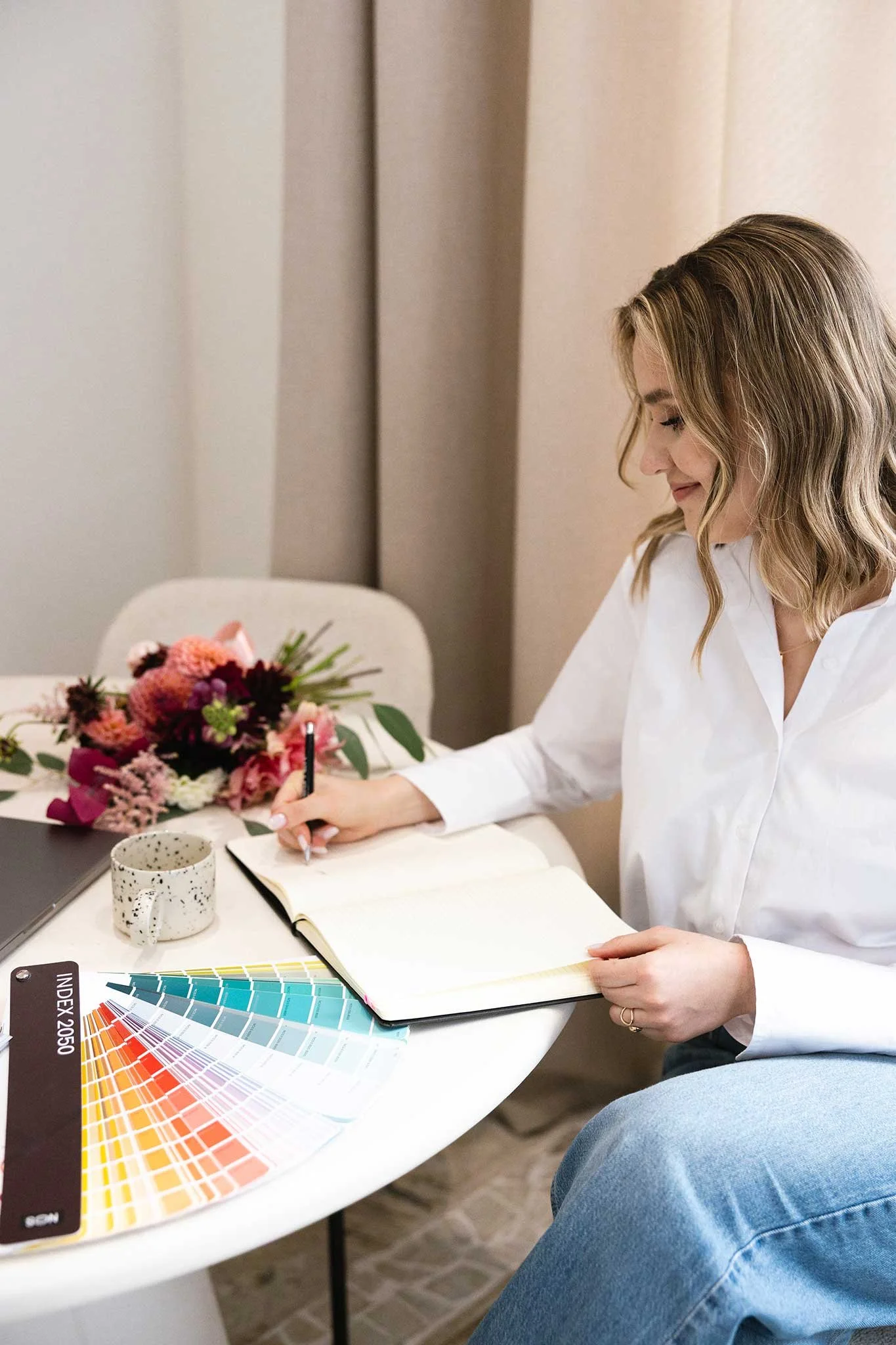 A woman with shoulder-length wavy blonde hair writing in a notebook at a table with a color swatch fan, a speckled mug, a laptop, and a bouquet of pink, red, and green flowers, indoors with beige curtains.