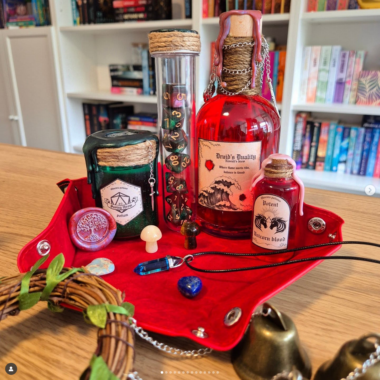 A red tray on a wooden table holds fantasy-themed items including potion bottles, charms, gemstones, a necklace, and small mushroom and mushroom-shaped objects, with a bookshelf in the background.