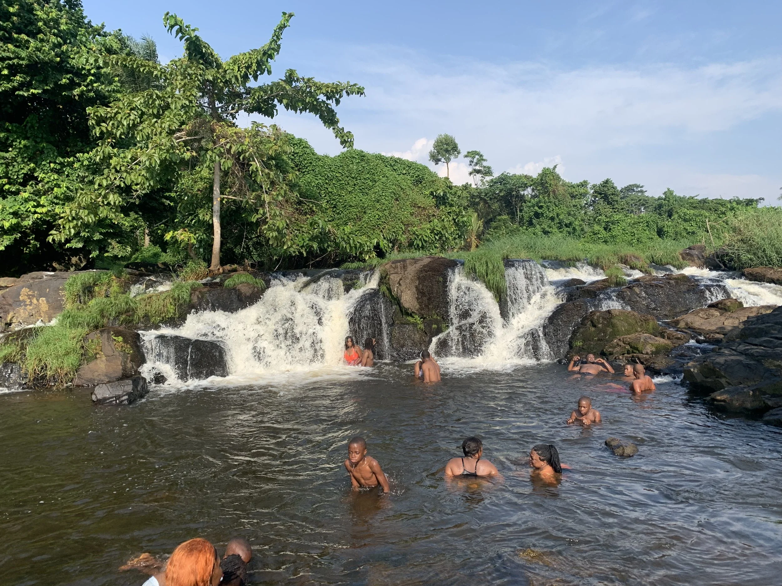 A group of people swimming and enjoying a small waterfall surrounded by lush greenery on a sunny day.