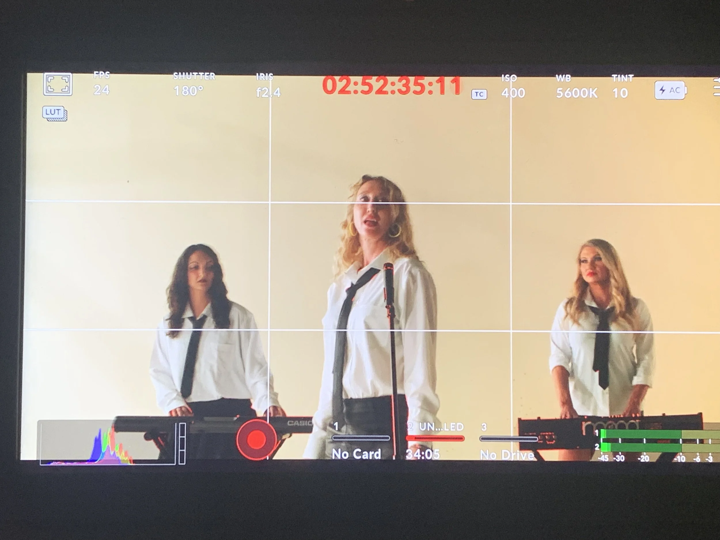 Three women in white shirts and black ties perform with keyboards, viewed through a camera screen.