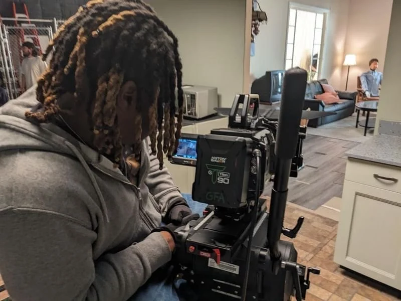 A person operating a professional film camera on a set with a living room scene in the background.