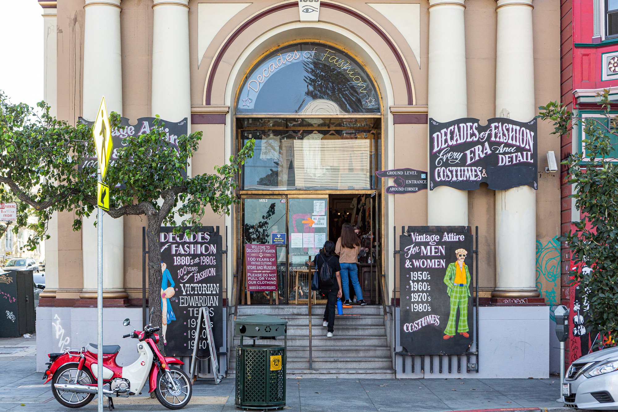 Entrance to a vintage clothing store with signs advertising fashion from the 1880s to 1980s, including costumes and vintage attire. A red and white scooter is parked near the entrance, and two people are entering the store.